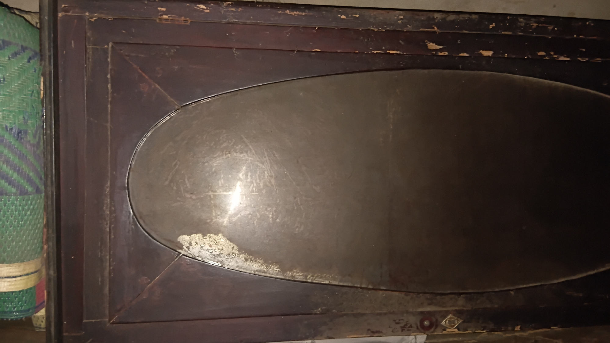 The image shows a close-up, eye-level view of an old, dark wooden cabinet with an oval-shaped, reflective surface in the center. The cabinet appears to be made of dark, possibly mahogany, wood with visible grain and some signs of wear and tear, including chipping paint and scratches. The oval surface is metallic, likely a mirror or polished metal, and is smudged and scratched, reflecting light from an unseen source. The top of the cabinet has a green, blue, and red woven mat placed on it. The setting appears to be an indoor environment, possibly a storage room or an older dwelling. There is no indication of the specific location within Prabumulih, Indonesia. The scene is static and depicts an object rather than an activity or interaction.