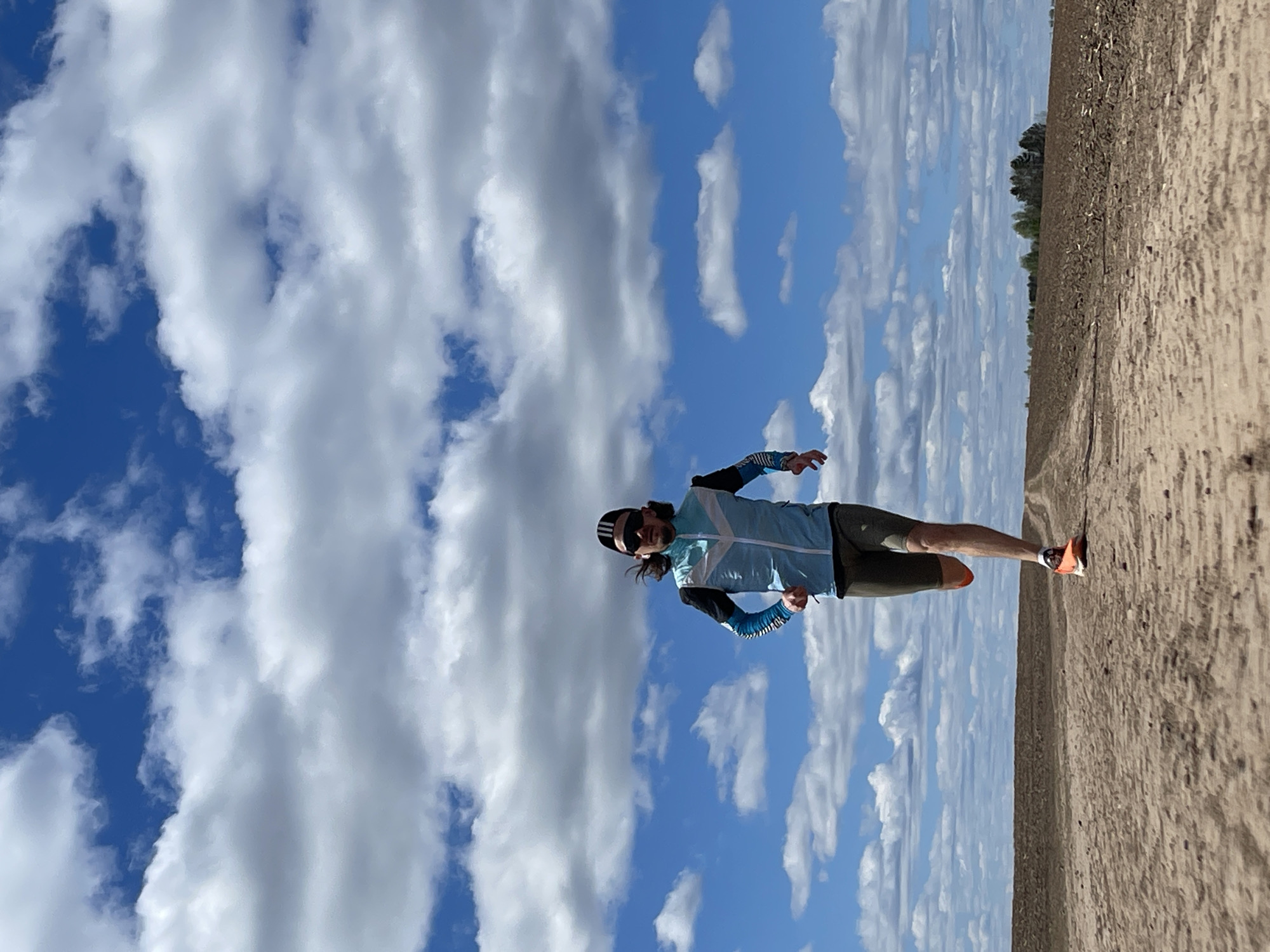 A runner, viewed at eye level, moves along a dirt path in Perevoznoe, Russia, on what appears to be a sunny day. The sky dominates the upper portion of the frame, filled with white, puffy clouds scattered against a blue backdrop. The man wears sunglasses, a blue and white cycling jacket, dark shorts, and orange shoes. The path is sandy and extends into the distance on the right side of the image, ending at a tree line. The photo shows the man mid-stride, suggesting action. The runner appears to be the only person in the scene.
