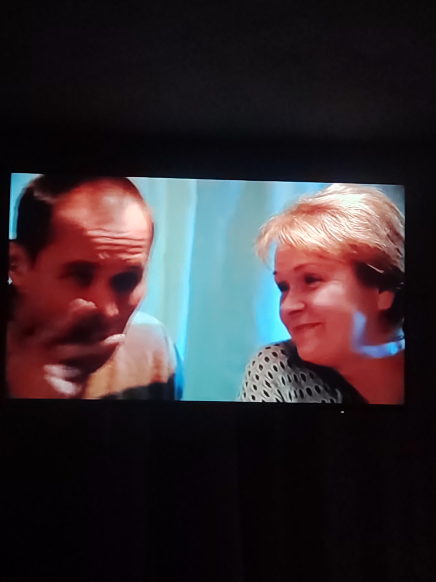 Two individuals, a man and a woman, are visible in this television screen capture. The man, on the left, has a bald head and is looking down and to the left, with his hand near his mouth. The woman, on the right, has short, blonde hair and is looking to her left and smiling. She is wearing a patterned top. The background appears to be a wall with blue and white striped curtains. The image is a screenshot from a television. The location of Chemuzhivka, Ukraine is not evident in this image.