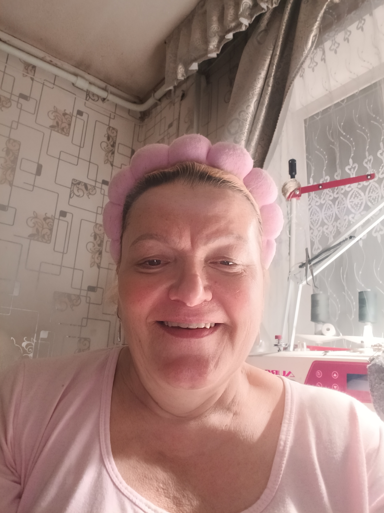 A woman with light skin and short blonde hair, wearing a pink, puffy headband and a light pink shirt, is smiling at the camera. She appears to be indoors, likely in a residential setting based on the wallpaper and curtain visible in the background. A sewing machine is also partially visible to the right, suggesting a crafting or sewing area. The location is Красный Маяк, Russia.