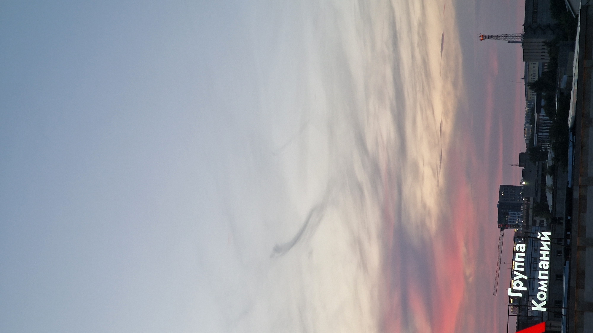 An image captures the sky over Kostanay, Kazakhstan, at dusk. The sky displays a gradient of colors, transitioning from a pale blue at the top to soft pinks and purples near the horizon, with wisps of clouds scattered throughout. In the foreground, illuminated signs are visible, displaying Russian text: "Группа Компаний" which translates to "Group of Companies". The signs are placed atop buildings, suggesting a commercial or industrial area. The silhouette of a construction crane and various low-rise buildings are discernible against the twilight sky. A tall, illuminated tower, likely a telecommunications mast, stands out to the right of the frame. The overall scene depicts an urban landscape bathed in the soft light of sunset or sunrise.