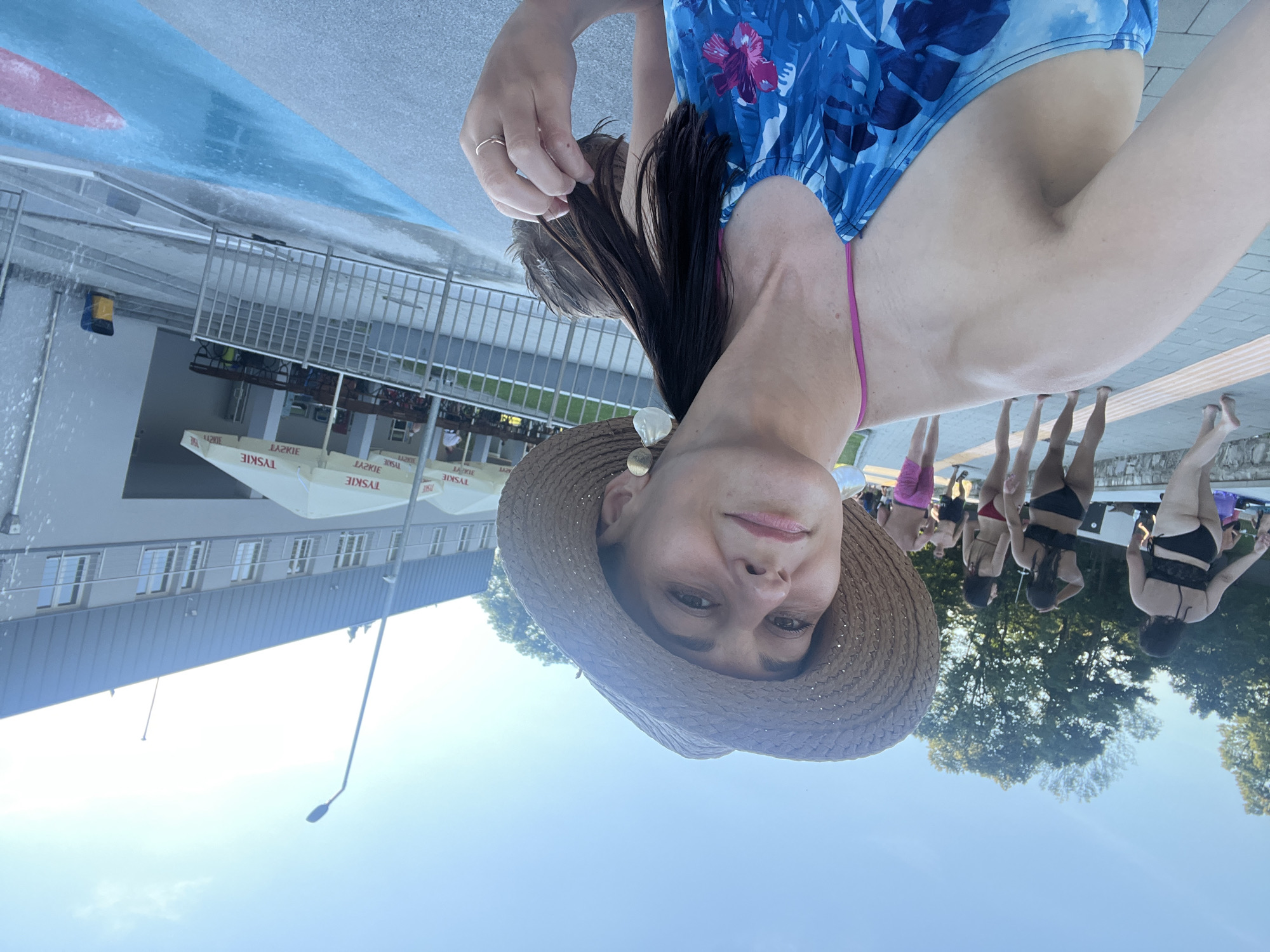 The image depicts a group of people at an outdoor pool. The primary subject is a woman in the foreground wearing a blue and white floral swimsuit and a straw hat. Her face is visible, and she appears to be taking a selfie. Her hand is raised, and she is touching her hair. Behind her, several other individuals are standing near the pool's edge, some in swimwear. The setting appears to be a sunny day, with clear blue skies. There are trees in the background, suggesting a natural or park-like environment. The pool area has a tiled edge and a blue and red painted section of the pool. Some white umbrellas with visible text are also present, with "TYSKIE" being legible on them. The overall mood seems relaxed and casual, indicative of a day spent at a swimming pool.