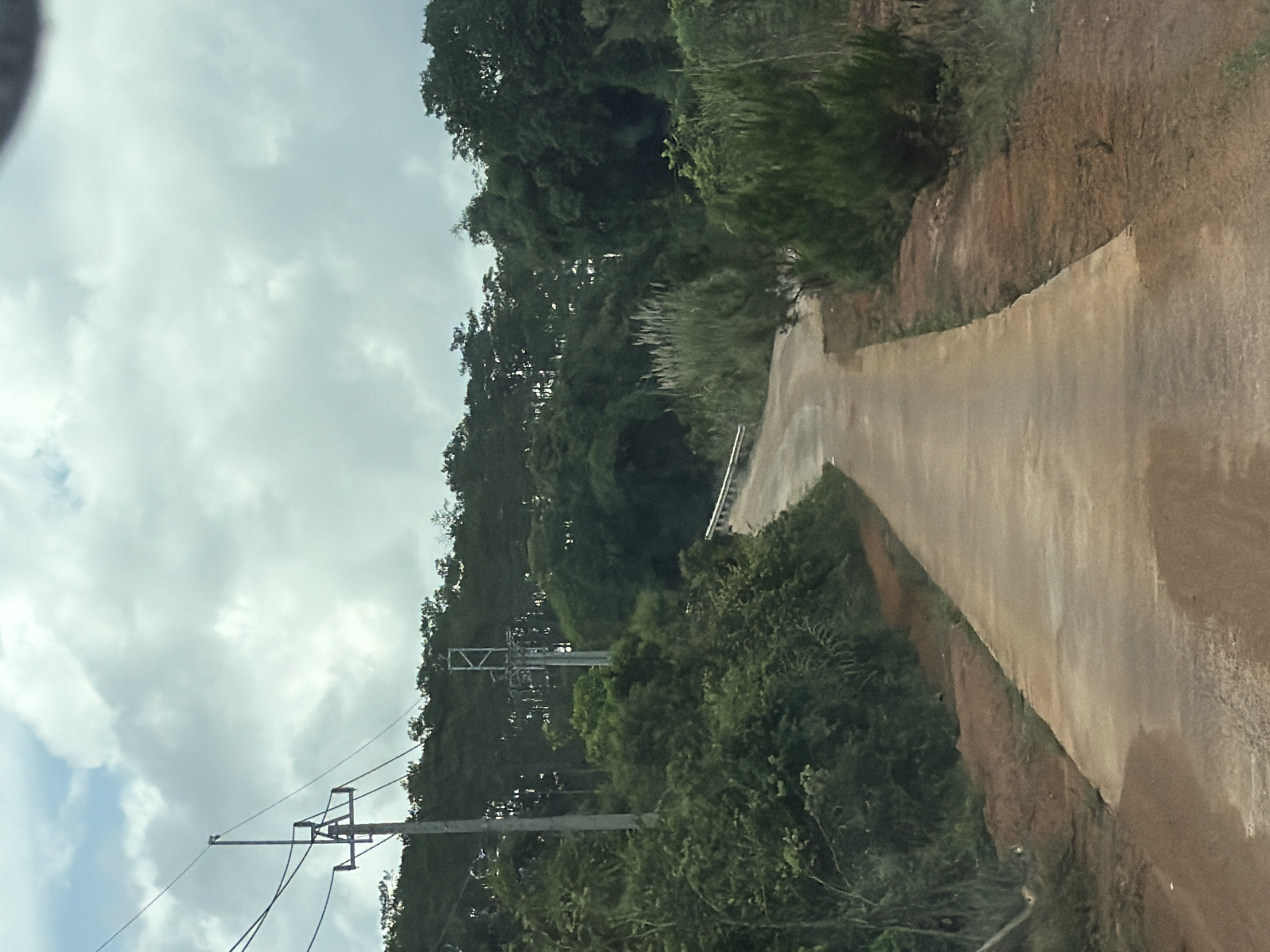 This media file, oriented incorrectly and depicting a scene from Măng Đen, Vietnam, features a narrow, paved road or path winding uphill through a verdant, hilly landscape. The road, appearing to be made of concrete or a light-colored material, shows damp patches, suggesting recent rain or high humidity. It curves gently to the left as it ascends into the distance.
Dense, lush green trees and varied vegetation, including taller trees and thick undergrowth, line both sides of the path, creating a rich, forested environment. On the immediate right, an earthen bank with exposed reddish-brown soil and scattered foliage borders the pavement, with a faint metal guardrail visible along the path's edge further up.
The sky is overcast with a blanket of grey and white clouds, indicating a cloudy daytime setting. In the foreground on the left, utility poles with multiple cross-arms and power lines are visible against the sky and the treeline, signifying the presence of infrastructure within this natural terrain. No people, animals, or specific activities are depicted. The scene conveys a sense of tranquility and remoteness, characteristic of a mountainous, forested region. There is no visible text in the image.