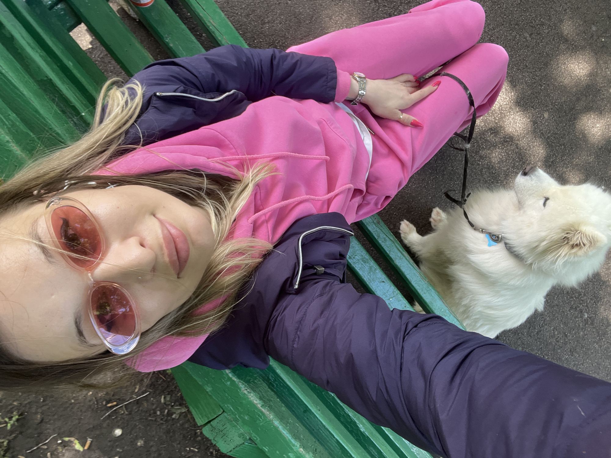 A person wearing pink sweatpants and a pink hoodie, a dark blue jacket, and pink-tinted sunglasses sits on a green park bench with a white dog lying at their feet. The person is looking up and smiling at the camera, holding a phone to take a selfie. The dog is facing away from the camera, with its head resting on the ground. The park bench is made of wood and painted green. The ground around the bench is paved with asphalt and littered with leaves and twigs. The image is taken from a high angle, looking down at the person and the dog. The overall scene suggests a casual outing in a park.