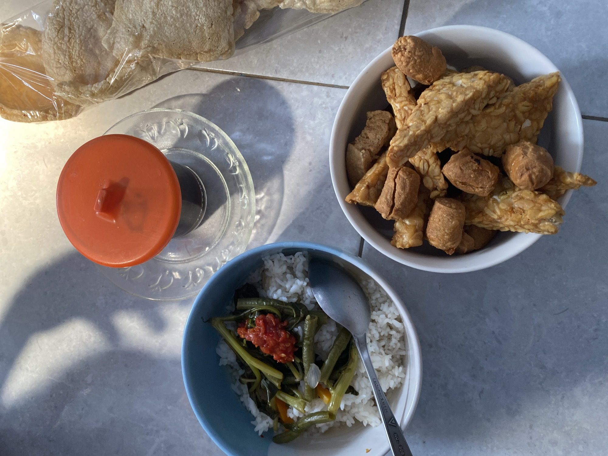 In Madiun, Indonesia, a meal is laid out on a tiled surface, bathed in dappled sunlight casting distinct shadows. The arrangement features three bowls. The first, a blue bowl, contains white rice topped with stir-fried vegetables, possibly water spinach and green beans, seasoned with a vibrant red chili paste. A metal spoon rests in this bowl. Adjacent to it, a white bowl is filled with fried tempeh pieces and what appear to be small, fried meatballs or nuggets. To the right, a clear plastic bag holds several pieces of pale, puffed crackers. In the background, a small glass with a dark liquid, likely coffee, is covered by an orange lid and placed on a glass saucer. The overall scene depicts a casual, home-style meal.
