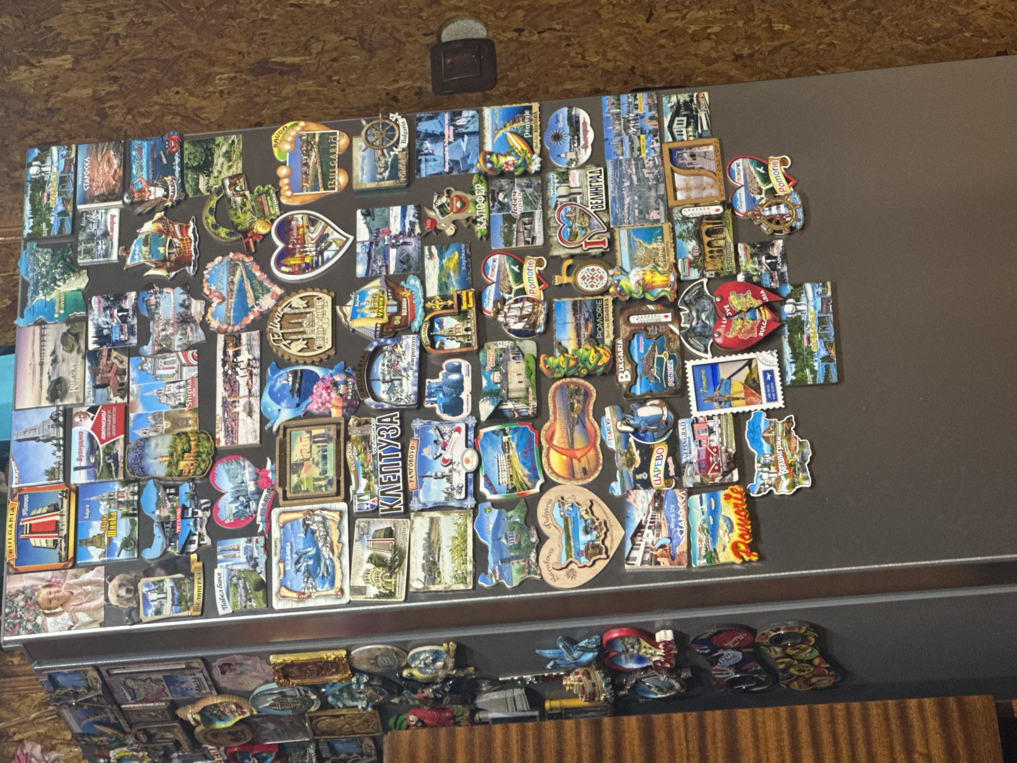 The image displays a refrigerator door densely covered with a vibrant collection of souvenir magnets. These magnets vary greatly in shape, size, and design, depicting a diverse range of subjects including landscapes, architectural landmarks, animals suchenses as a bear and a dolphin, traditional boats, and abstract patterns.
A significant amount of text is visible on the magnets, predominantly in both Cyrillic and Latin scripts. Many magnets explicitly state "BULGARIA" and feature names of various Bulgarian cities and regions, such as "Velingrad" (ВЕЛИНГРАД, Велинград), "Pomorie" (ПОМОРИЕ, FOMORIE, Pomorie, Demorie), "Pamporovo," "Shipka" (Шнока), "Tsarevo" (ЦАРЕВО, TSAREVO), "Burgas," "Pleven," "Tryavna," "Pavel Banya," "Lovech," "Kardzhali," and "Starosel" (СТАРОСЕЛ). Other texts include phrases like "Wine & SPA," "Thirsty," and "КОМУНИЗМЪТ" (Communism). One prominent magnet features a young girl.
The background wall above the refrigerator is textured, possibly wood paneling. A small, dark, rectangular object with a silver top is attached to the top-center of the refrigerator door, above the main magnet cluster. Below the main collection, a shelf is visible, holding additional small items, some of which appear to be more magnets or decorative objects, against a wooden surface. The extensive collection of Bulgarian-themed magnets strongly indicates the setting is in or related to Bulgaria, aligning with the provided context of Sarnevo, Bulgaria, and reflects a personal history of travel and souvenir collecting.