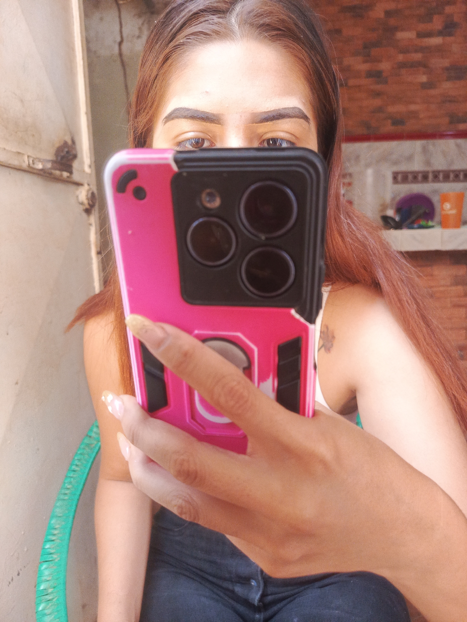 A fair-skinned woman is taking a selfie with her phone, which has a pink case. She has brown hair and is wearing a white top and dark jeans. The background features a brick wall with a shelf that has various items on it, as well as a white wall with an open metal door. The woman appears to be sitting on a green chair. No specific indicators in the image confirm the location as Juan Griego, Venezuela, but the environment suggests an indoor or semi-outdoor space.