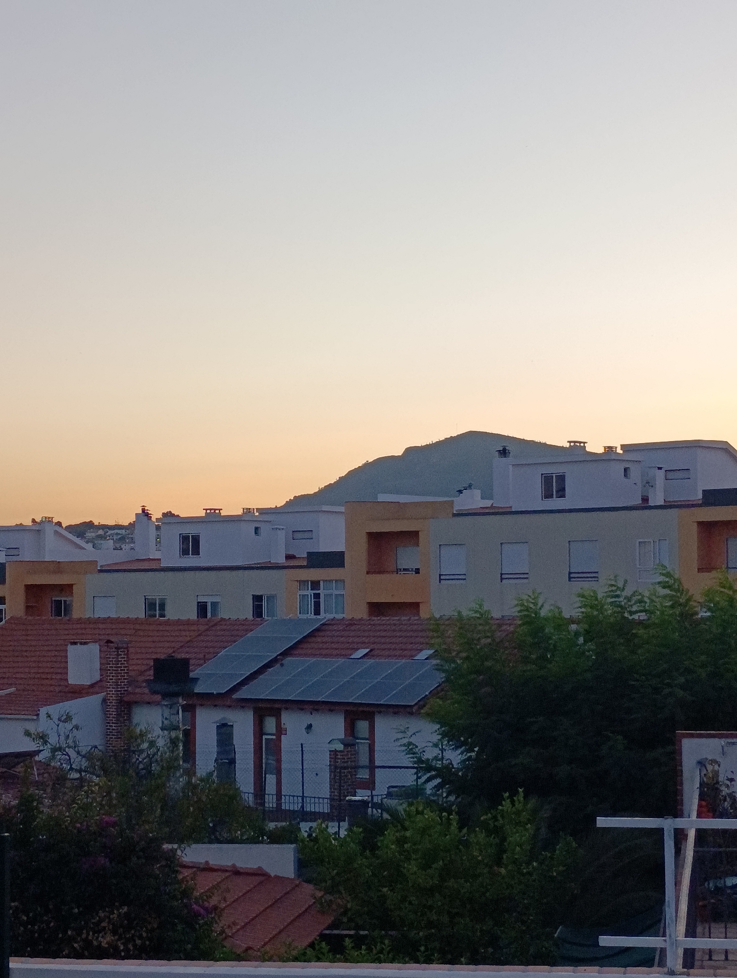 This is a medium-shot, eye-level, vertical photograph taken from a slightly elevated position, looking out over a residential area in Setúbal, Portugal, during a soft sunset. The sky is a gradient of pale yellow at the horizon, transitioning to a light blue at the top. A distant, rounded, green-covered hill dominates the background. In the mid-ground, a cluster of multi-story buildings with flat roofs are visible. The buildings have varying facade colors, including light yellow, white, and peach, with numerous windows, some of which appear to be covered by shutters. Several buildings have prominent white air conditioning units or vents. Red-tiled roofs are visible on some of the lower buildings in the foreground. Solar panels are installed on the roofs of at least two buildings. Dense green foliage, including trees and bushes, partially obscures the lower portions of the buildings and create a natural foreground element. There are no people visible in the image. The overall scene depicts a peaceful, urban residential landscape under a warm evening sky.