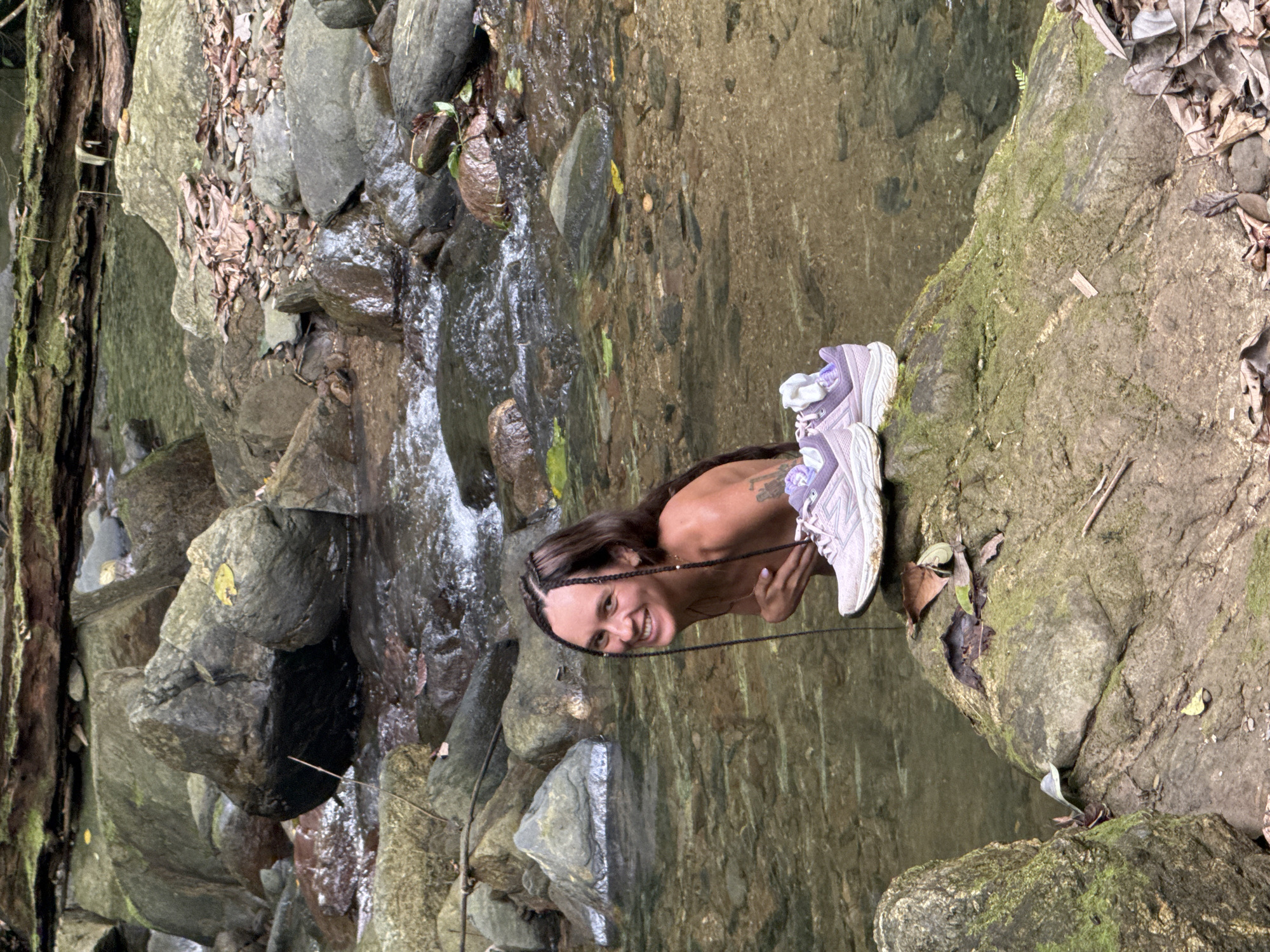 A woman is positioned on a rock outcrop next to a stream, possibly in Suối Giàng, Vietnam. She is smiling and looking directly at the camera. She has long brown hair styled with braids. Next to her are a pair of lavender-colored athletic shoes. The stream flows over rocks and through a lush, green environment. The scene is natural and appears to be outdoors.