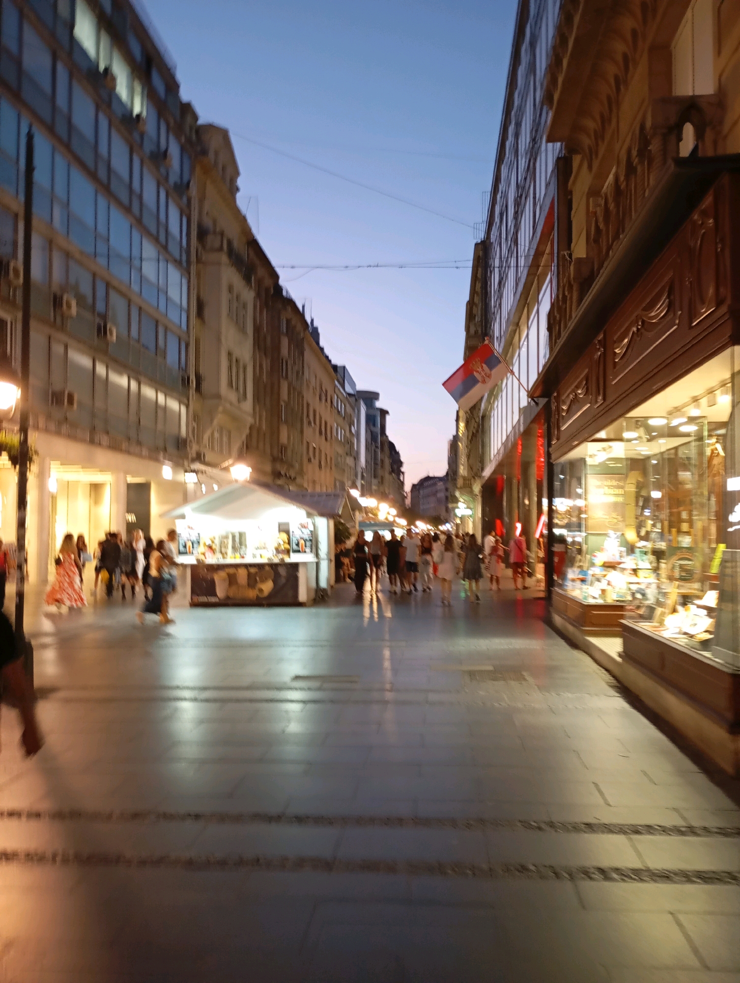 The image depicts Knez Mihailova Street in Belgrade, Serbia, at dusk. The street is lined with tall buildings, featuring a mix of modern and older architecture. On the left, a glass-fronted building with multiple stories dominates the foreground. On the right, a traditional building with ornate decorative elements and a large storefront display is visible.
A Serbian flag flies from a pole on the building on the right. Pedestrians are walking along the street, some blurred due to motion. Several streetlights are illuminated, casting a warm glow. In the middle ground, there is a white tent-like structure, possibly a kiosk or a temporary stand, with people interacting around it. The sky is a deep blue, indicating the transition from day to night. The street surface is paved with large tiles, and there are decorative stone inlays. The overall scene suggests a lively urban environment in the evening.