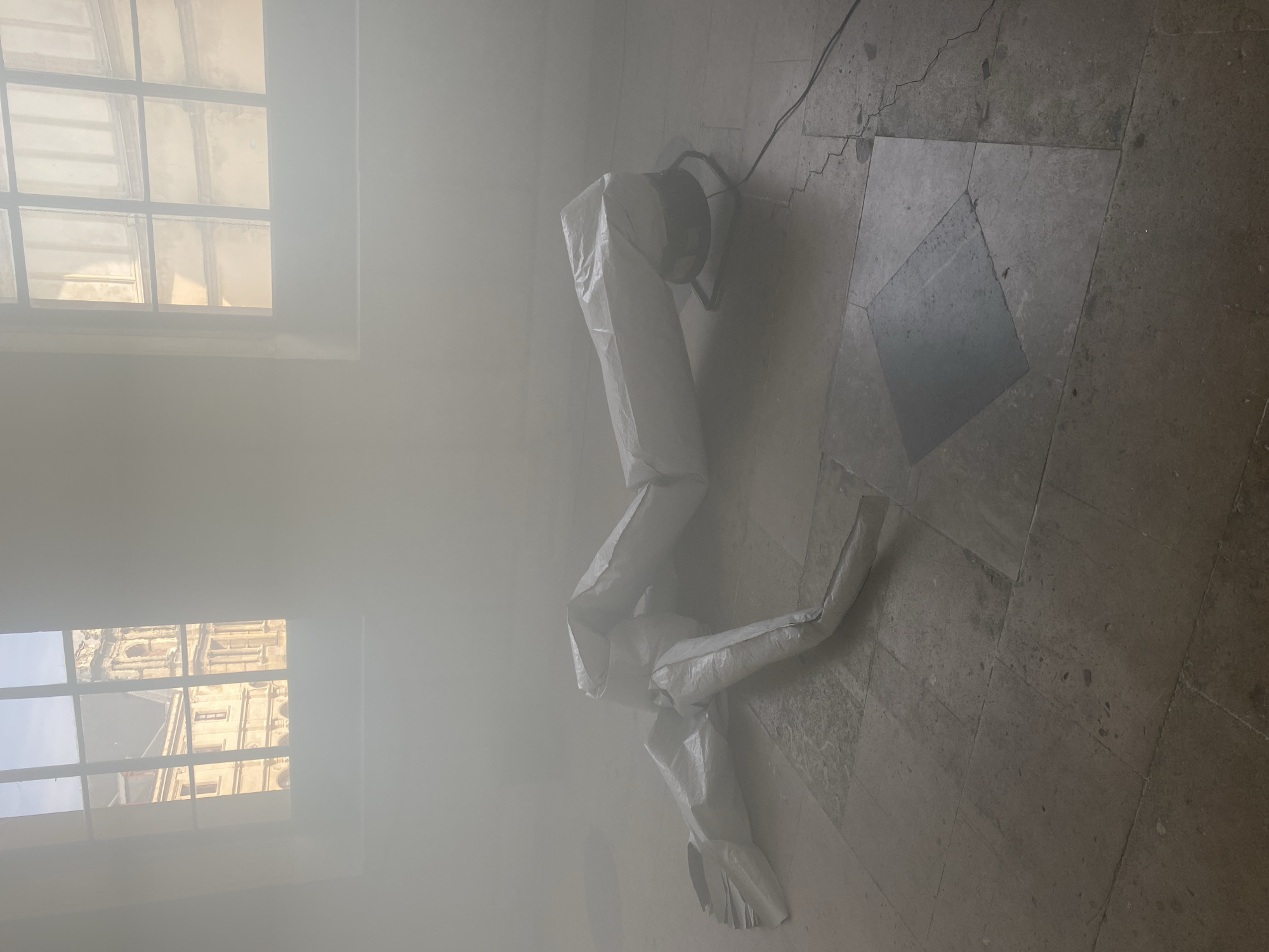 The interior of a building in Paris, France, is filled with a heavy fog. Two windows are visible, one showing a building exterior. The floor is tiled with large stone slabs. An inflated white object, resembling a mannequin or a tube, lies on the floor, connected to a fan. The air is still and visibility is low due to the dense fog.
