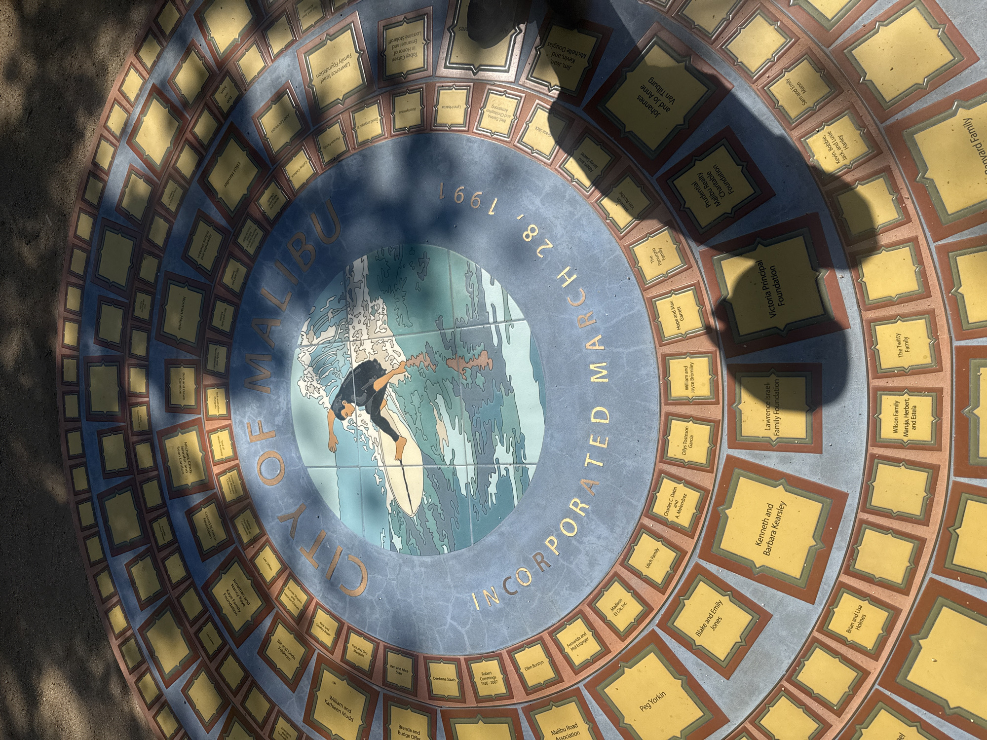 This is an image of a circular mosaic featuring a surfer riding a wave at its center. Encircling the surfer image are the words "City of Malibu Incorporated March 28, 1991". Around this are rings of small rectangular plaques bearing names and affiliations, possibly donors or significant figures associated with the city. The mosaic is likely located in Malibu, California, given the text and imagery. The lighting suggests it is outdoors and during the daytime.