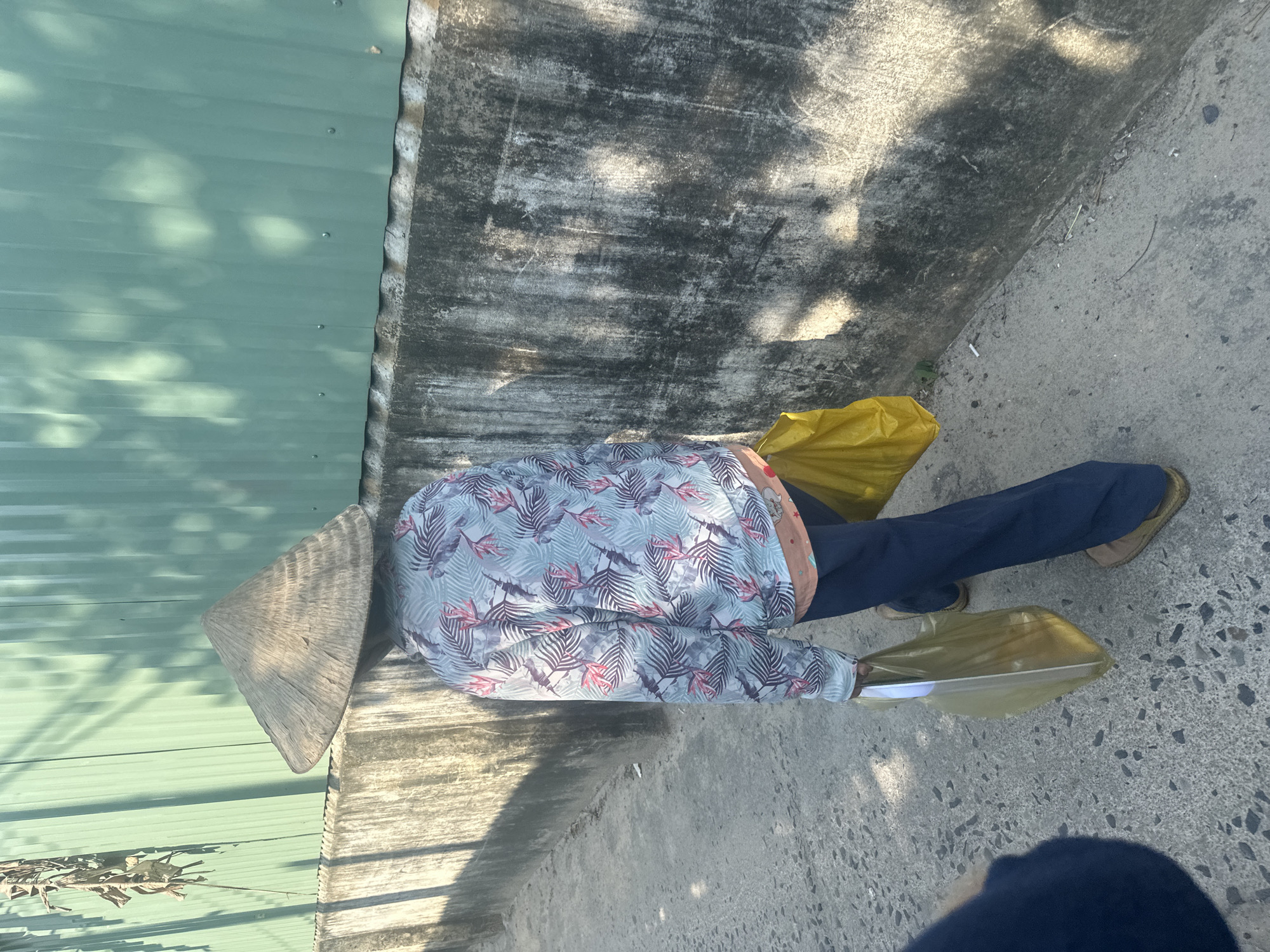 A person wearing a conical hat, a light blue leaf-patterned shirt, and dark blue pants is lying on the pavement next to a green corrugated metal wall and a weathered concrete wall in Bac Nha Trang Ward, Vietnam. The person is partially obscured by a yellow plastic bag, and holds another plastic bag. The pavement is grey and concrete. Shadows are cast on the walls.