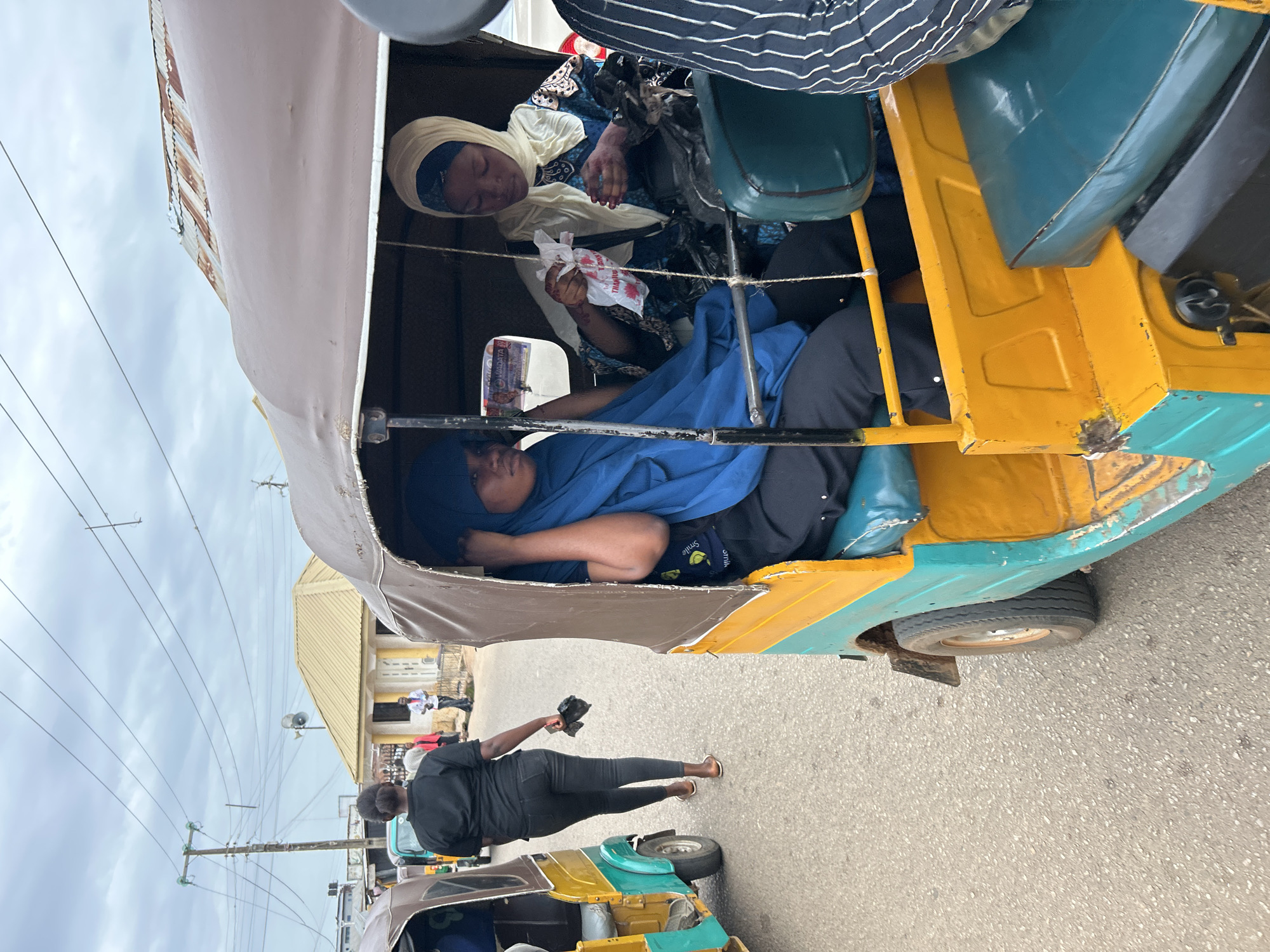 Two women are seated inside a yellow and green auto-rickshaw in Jos, Nigeria. The woman on the left is wearing a blue hijab and dark clothing. The woman on the right is wearing a white hijab and a blue patterned outfit. She is holding a small white plastic bag. In the background, a woman in a black shirt and black pants is walking away from the camera, carrying a dark plastic bag. Other auto-rickshaws are visible, along with a building with a yellow roof and power lines overhead. The scene appears to be a street in a city.