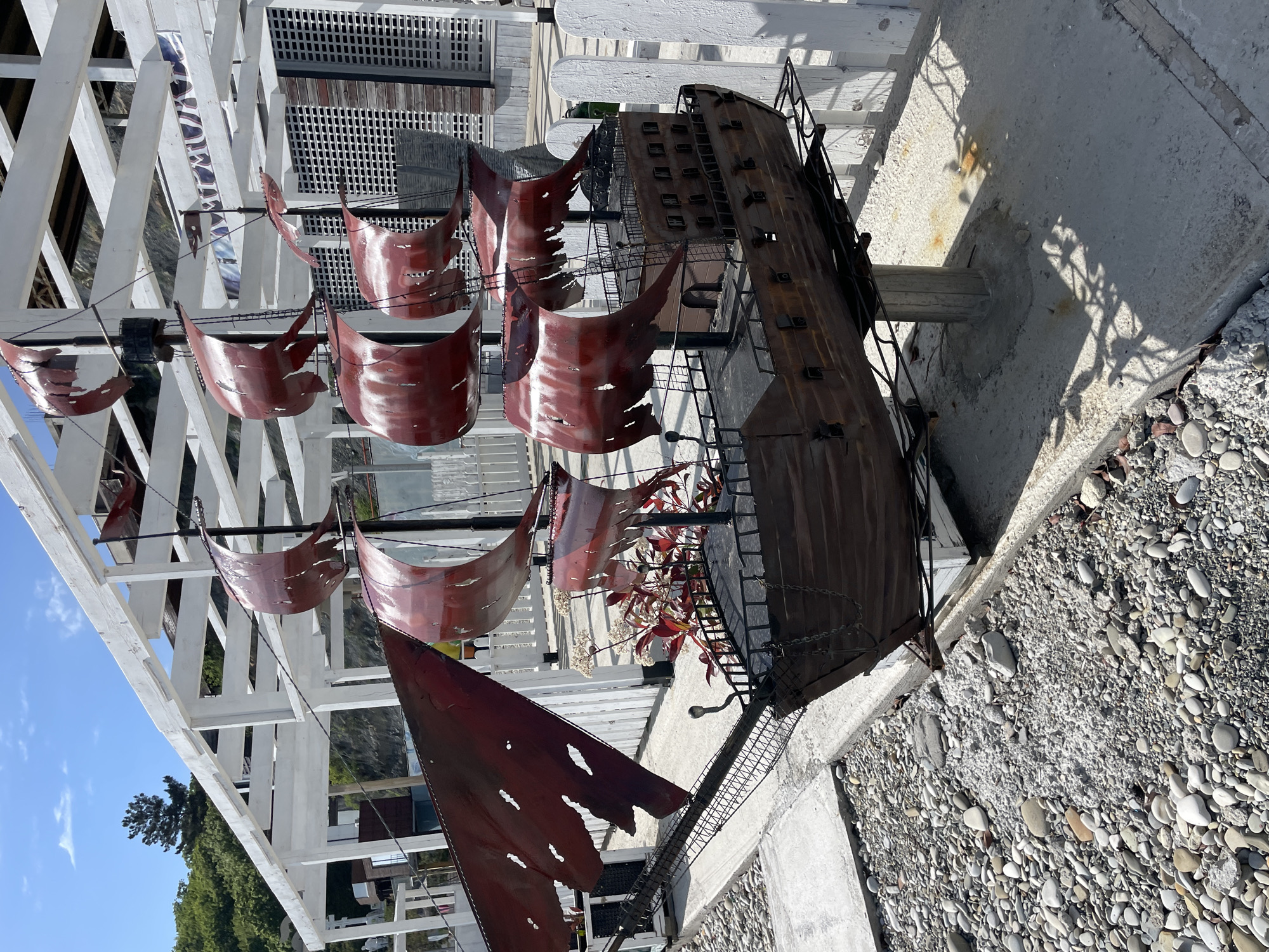 This media file presents a large, decorative metal sculpture of a historic sailing ship, likely a galleon or pirate vessel, situated outdoors under bright daylight in Olginka, Russia. The ship model features a dark, weathered metal hull with details resembling portholes or windows, and intricate metal railings. Its sails are made of reddish-brown metal, intentionally sculpted to appear tattered and aged, contributing to a rugged, old-world aesthetic.
The sculpture rests on a base composed of concrete and an area covered with smooth, light-colored pebbles, reminiscent of a beach or riverbed. Surrounding the ship is a distinctive white wooden trellis or lattice structure, possibly part of a pergola or building facade, which casts sharp shadows on the ground due to the clear, sunny conditions. Behind the ship, glimpses of green and reddish foliage with small white flowers are visible, along with distant green trees and a clear blue sky dotted with a few clouds.
A banner or sign, partially visible hanging from the white structure behind the ship's sails, displays the text "ОЛУМПИЯ" (Olympia). This suggests the ship is part of a themed display for a local business or establishment. The scene is static, with no human activity, focusing solely on the elaborate ship sculpture and its immediate surroundings. The overall impression is one of a charming and robust outdoor art installation designed for visual appeal.