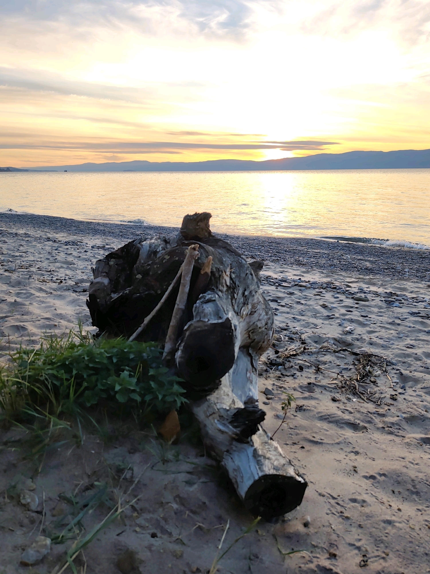 A log rests on a sandy beach next to a body of water, with mountains in the distance. The sky is filled with clouds and the sun is setting. The scene appears to be in Kharantsy, Russia.