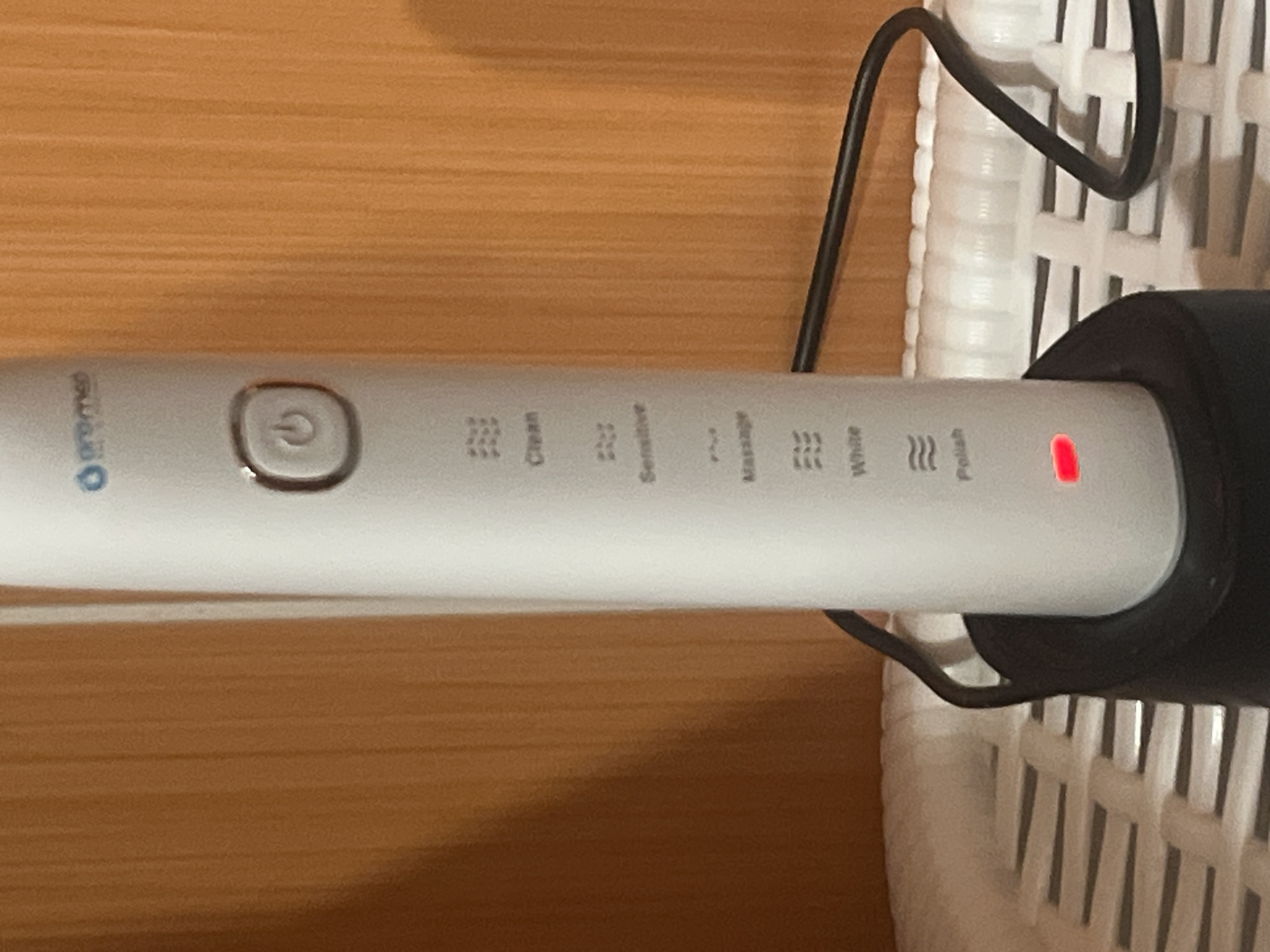 The image shows a close-up of a white electric toothbrush in its charging base. The toothbrush has a circular power button with a power symbol on it. Below the power button are several modes indicated by icons and text: "Clean", "Sensitive", "Massage", "White", and "Polish". A small red light is illuminated on the toothbrush, possibly indicating it is charging or in a specific mode. The toothbrush is resting in a black charging base, which is plugged in by a black cord that goes out of frame. The toothbrush is positioned on a white textured surface, possibly a shelf or countertop. The background is a wooden surface with vertical grain, likely a wall or furniture. The overall lighting is dim, with a focus on the toothbrush. There are no people or animals visible in the image. The scene is indoors. This image is not from Rybnitsa, Moldova.