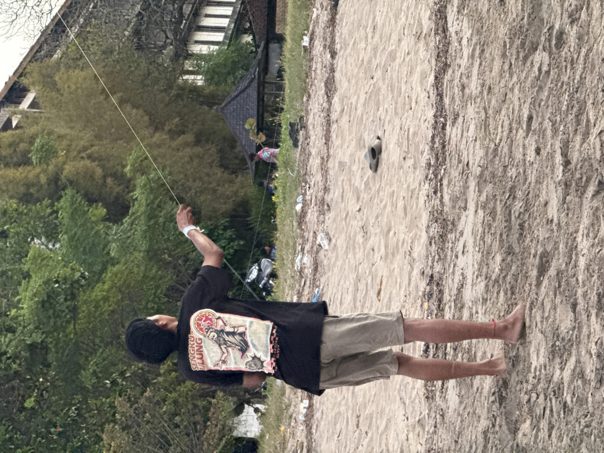 A person stands on a sandy beach, holding a kite string. The person is wearing a black t-shirt with a graphic design and text that appears to say "SENGKU-ULUNG", and light-colored shorts. The person's right arm is raised, holding the kite string that extends upwards and out of frame. The beach is covered in sand, with some debris scattered on the surface. In the background, there are trees and a building with a dark-tiled roof. Other people are visible in the distance, and there is a dark object on the sand that resembles a shoe or sandal. The lighting suggests it is daytime, possibly overcast. The scene appears to be in Kuta Selatan, Indonesia, a location known for its beaches.