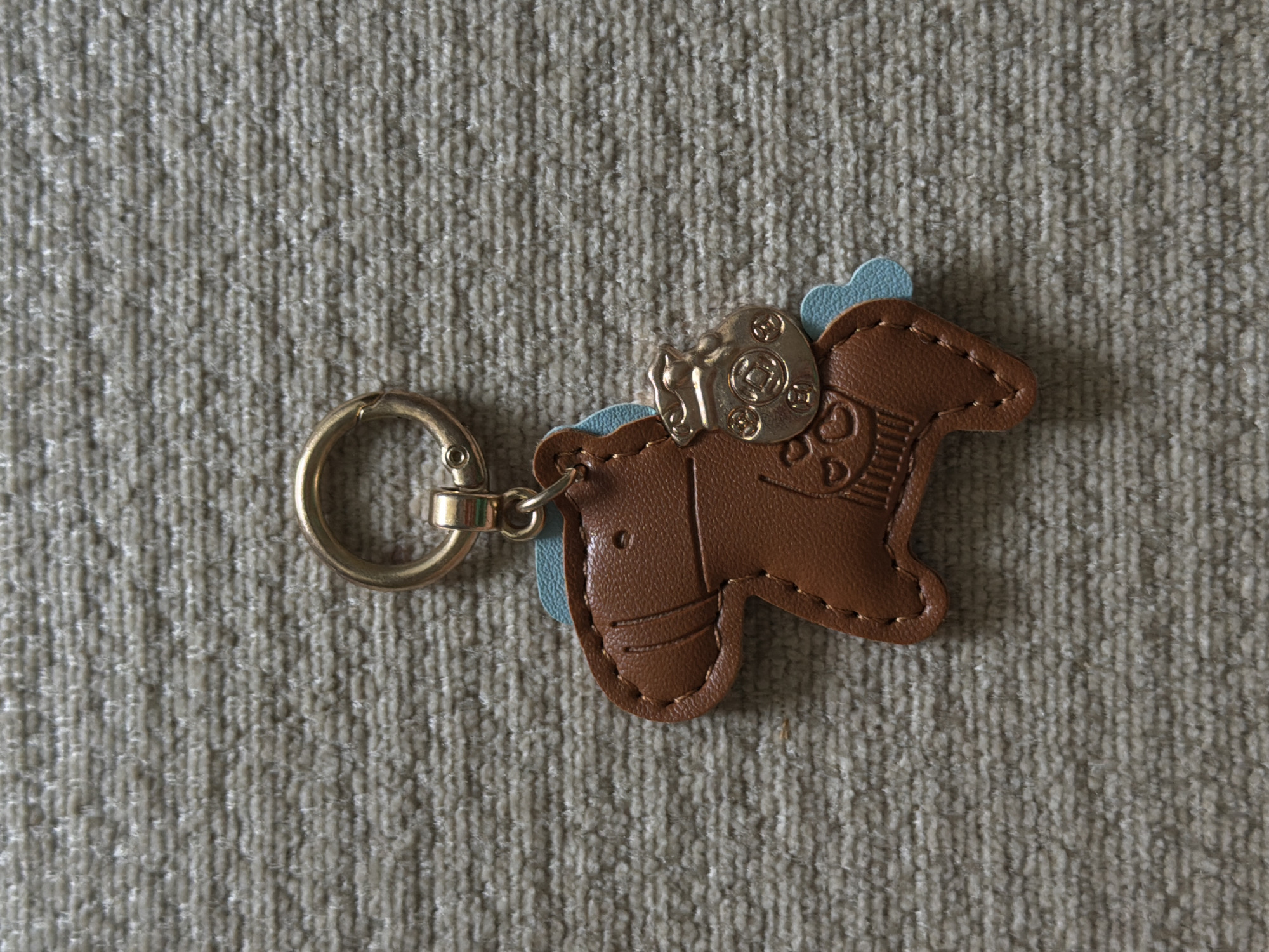 A brown leather keychain in the shape of a horse with light blue accents is lying on a beige, textured surface. The horse has stitched details to represent its mane, eyes, and legs. Attached to the horse's back is a small, gold-colored charm shaped like a money bag. A gold-colored ring is connected to the top of the horse via a small clasp. The scene appears to be an overhead shot of the keychain on a carpet or fabric surface. There is no discernible location within the city of Chirchik, Uzbekistan.