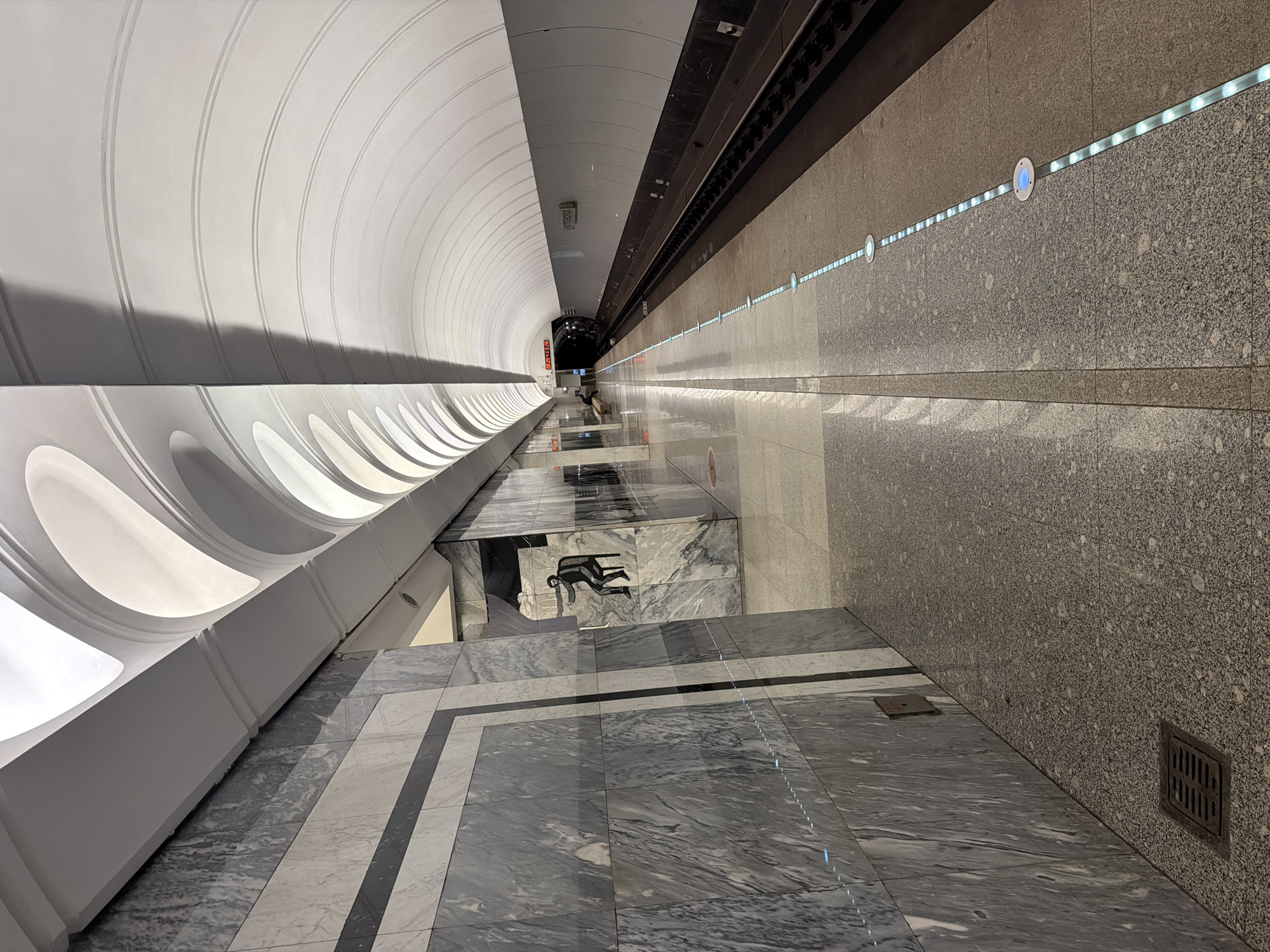 An empty subway platform in Moscow, Russia features a vaulted ceiling with intricate white arches that create a dramatic play of light and shadow. The walls are clad in polished grey marble with darker veins, and a stylized black silhouette of a seated figure is visible on one section of the wall. The platform floor is a speckled grey granite. A dark subway track runs along the right side of the platform, bordered by a line of blue illuminated circular markers. In the distance, a digital clock displays "03:20:44:26". The overall scene conveys a sense of modern architectural design and emptiness.