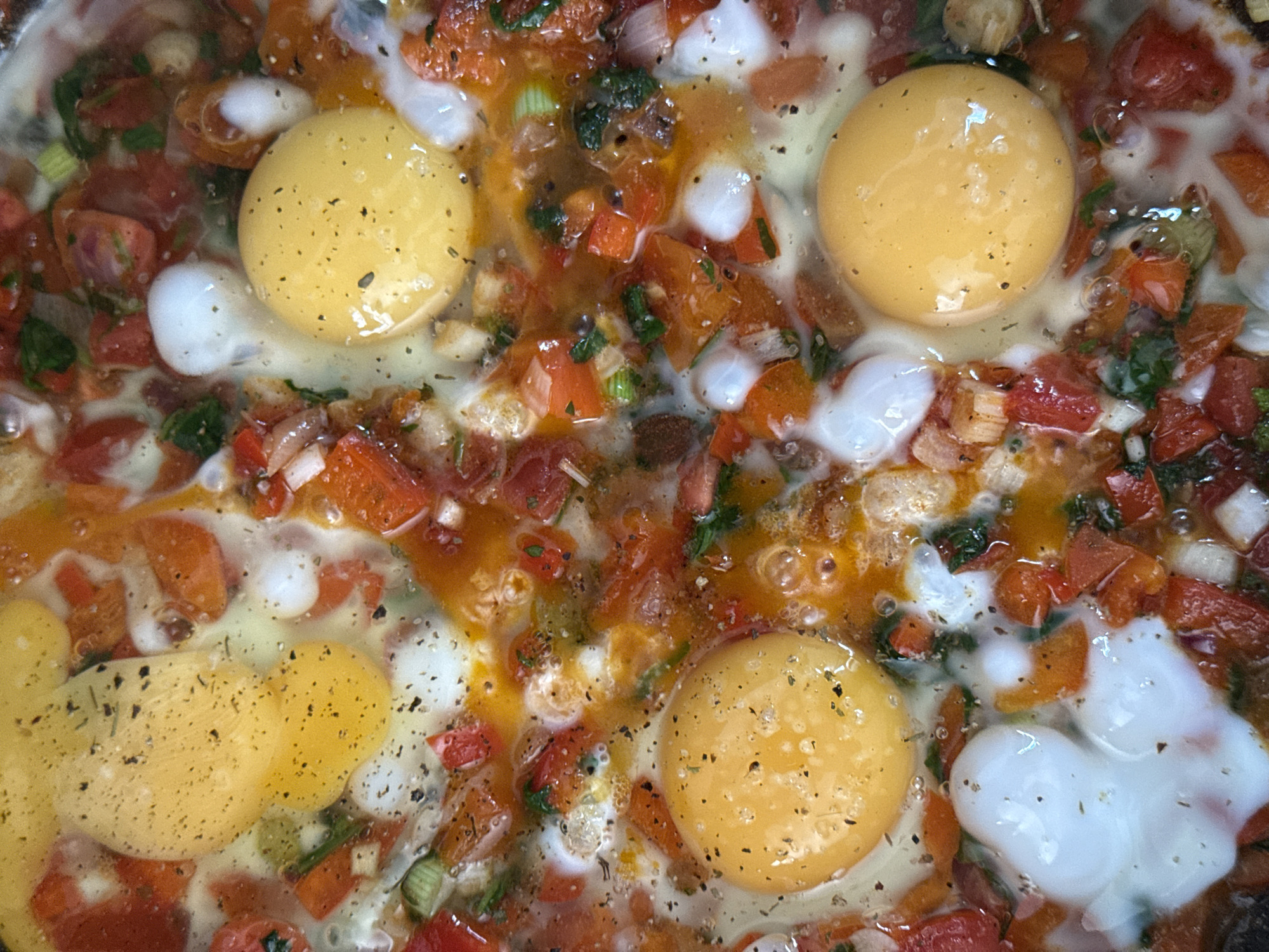 This is a close-up, overhead shot of a dish being cooked, likely a shakshuka or a similar egg-based meal. The dish contains several whole eggs, with visible yolks, cracked into a mixture of chopped tomatoes, red peppers, onions, and herbs. The ingredients are submerged in a liquid, likely tomato sauce, and the dish is seasoned with black pepper. The scene is shot from directly above, with no context of its location, people, or environment. Therefore, its location in the city of Chaikovsky, Russia, cannot be determined from the image alone.