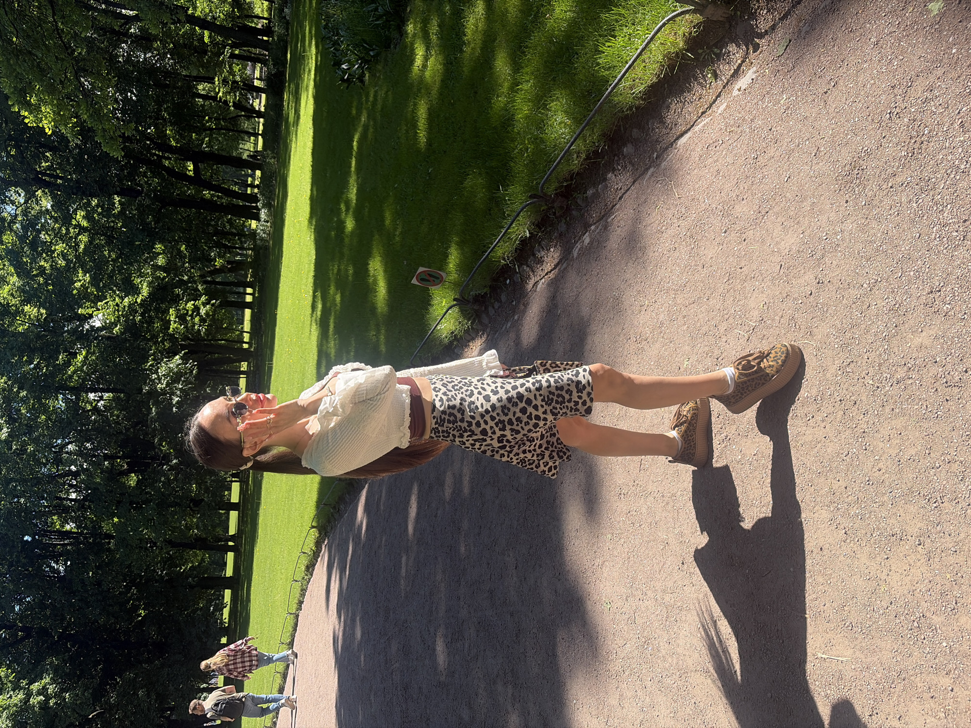 The image captures a sunlit scene in a park within Saint Petersburg, Russia, on a clear day. In the foreground, positioned on the right, a young woman stands on a light brown gravel path. She has long brown hair tied back with a light-colored scrunchie and wears round sunglasses. Her attire consists of a ruffled, off-white long-sleeved crop top over a dark maroon bralette, a leopard-print mini-skirt, and matching leopard-print platform sneakers with brown laces. She has multiple rings on her right hand. Her left hand is raised to her face, touching her chin near her sunglasses, while her right hand holds a small leopard-print bag or clutch. Her body is angled slightly away from the camera, and she is looking to her right (viewer's left).
Behind her, to the left of the path, two other individuals walk away from the camera. The person on the left is a male wearing a light shirt, blue jeans, and a dark backpack. The person beside him is a female in a plaid shirt and blue jeans.
The path is bordered on both sides by lush green grass, which is separated from the path by a low, dark metal railing. A small, round sign with a red circle and a diagonal line, possibly indicating a prohibition, is visible near the railing on the right side of the path. The background features a dense line of tall, mature trees with abundant green foliage, indicative of a forest or large parkland. Strong sunlight from the left casts long, distinct shadows of the people and trees across the gravel path and the grass, creating areas of bright illumination and deep shade.
