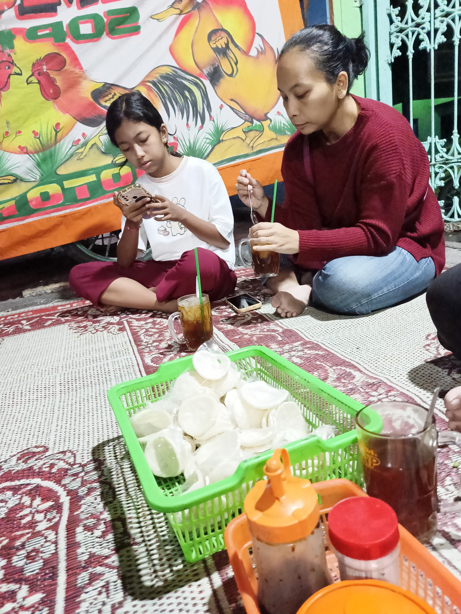 In Kebondalem Kidul, Indonesia, two individuals are seated on a woven mat outside at dusk. A young girl, wearing a white t-shirt and maroon pants, is engrossed in her mobile phone. Beside her, an adult woman in a maroon sweater and blue jeans holds a glass of iced tea, using a fork to stir it. In the foreground, a green plastic basket overflows with white crackers, and an orange plastic tray holds bottles of sauce and a jar with a red lid. A banner depicting roosters and the numbers "402" is visible in the background, along with a green metal fence. The scene suggests a casual outdoor gathering or meal.