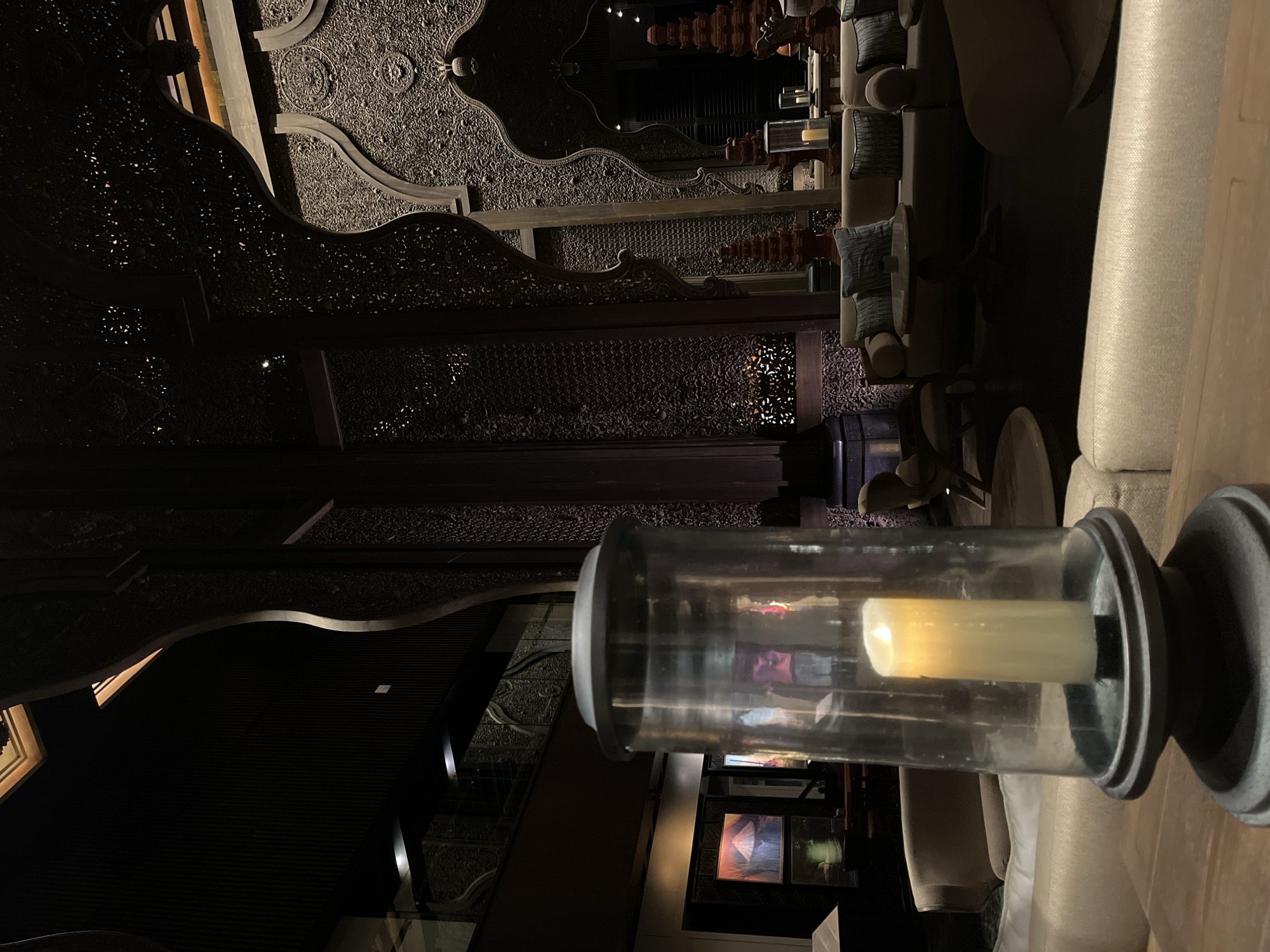 An indoor scene depicts a dimly lit, luxurious lounge area. In the foreground, a glass lantern with a lit white candle sits on a wooden table. Behind the lantern, plush sofas and armchairs are arranged around low tables, suggesting a comfortable seating area. The background reveals an ornate, dark wooden interior with intricate carvings on the walls and arches, creating a sophisticated and perhaps Middle Eastern or Asian-inspired ambiance. Paintings are visible on a wall to the left. The overall lighting is subdued, with highlights from the candle and other ambient sources. The location is identified as Kuta Selatan, Indonesia.