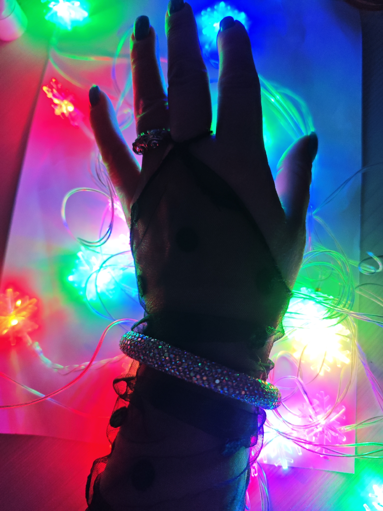 A hand wearing a black, sheer polka-dot glove and a sparkling bracelet is positioned on a white surface surrounded by colorful LED lights. A ring adorns one of the fingers. The lighting is predominantly red, green, and blue. The location is not discernible from the image, and there is no indication that the image was taken in Markovskii, Russia.
