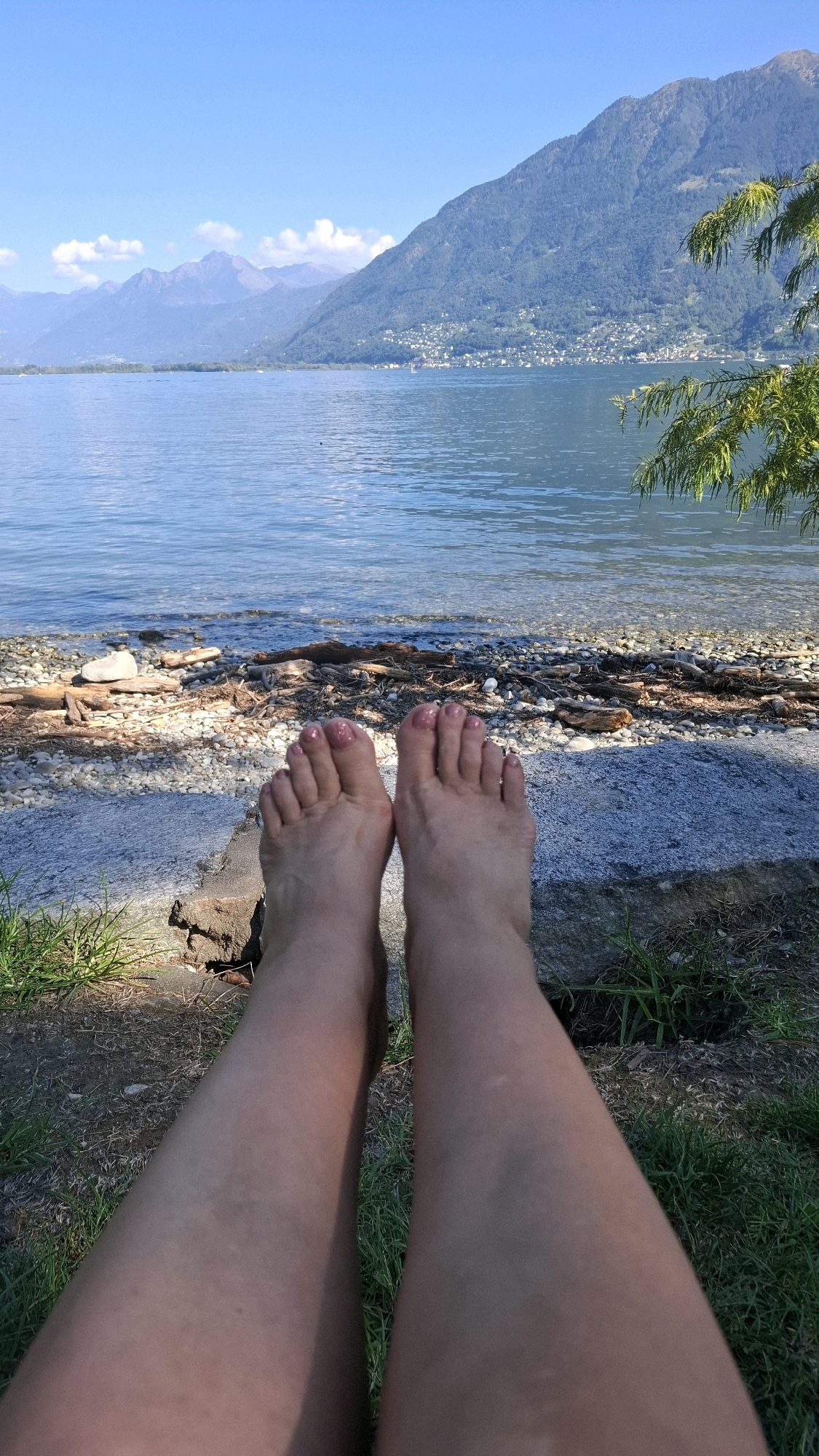 A close-up shot of a person's bare legs and feet rests in the foreground. The feet are positioned centrally, with toenails painted a shimmery pink. Beyond the feet, a rocky shoreline leads to a calm, blue lake. In the distance, a verdant mountain slopes upward, dotted with buildings that form a town. The sky above is a clear, bright blue. The overall impression is a peaceful scene at the edge of a lake, likely in or near Locarno, Switzerland, given the characteristic landscape of the region.