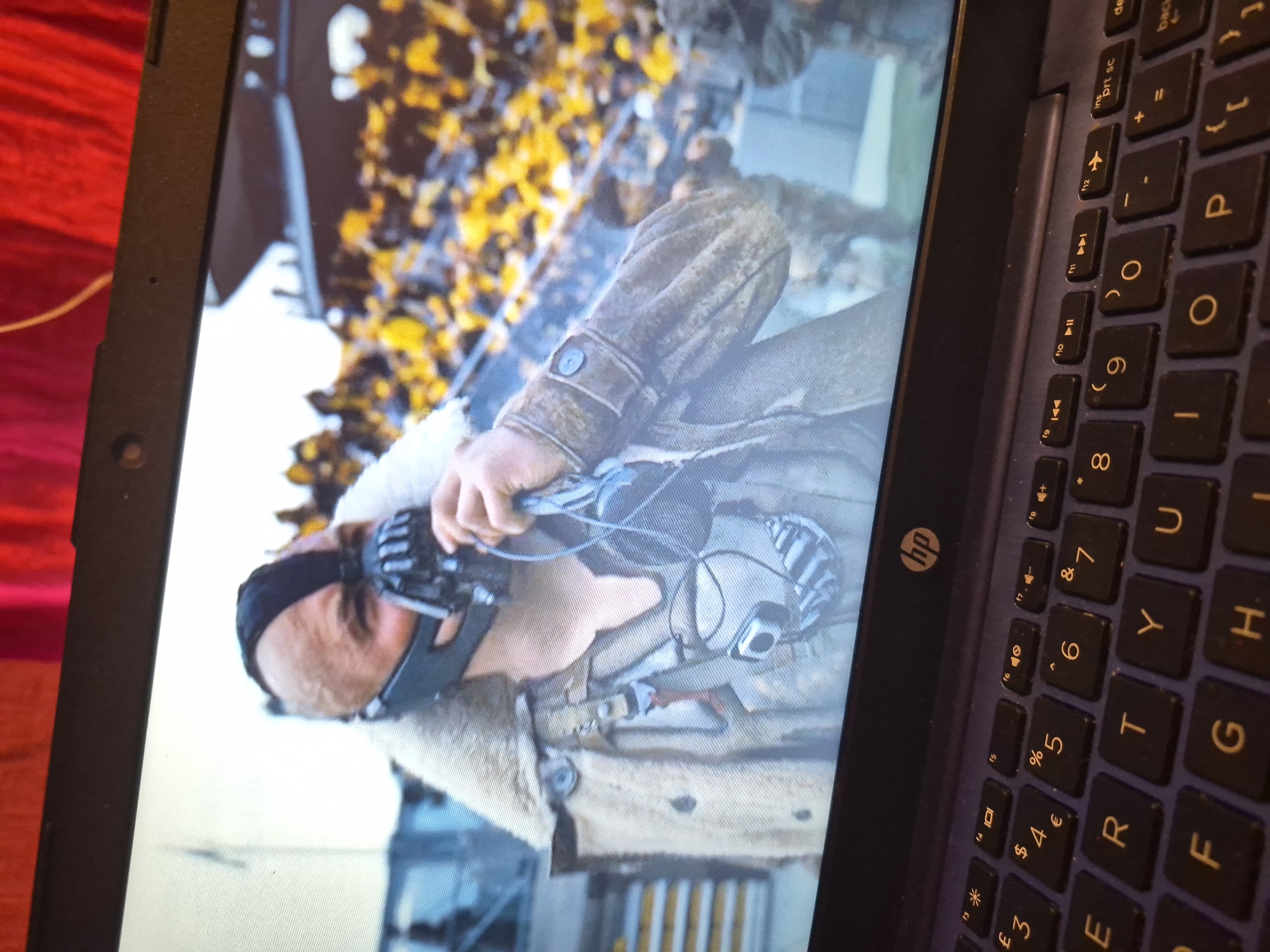 A close-up, angled shot shows a laptop screen displaying a movie scene. On the screen, a man wearing a distinctive mask and a fur-lined coat is visible, holding a device to his ear. He appears to be on a field or stadium, with a crowd of people in the background wearing yellow attire. The laptop keyboard is in the foreground, with the HP logo visible. The overall image is a photograph of a laptop screen displaying a scene from a film. There is no information in this image that would allow for the determination of a location in the city of Sandwell, United Kingdom.