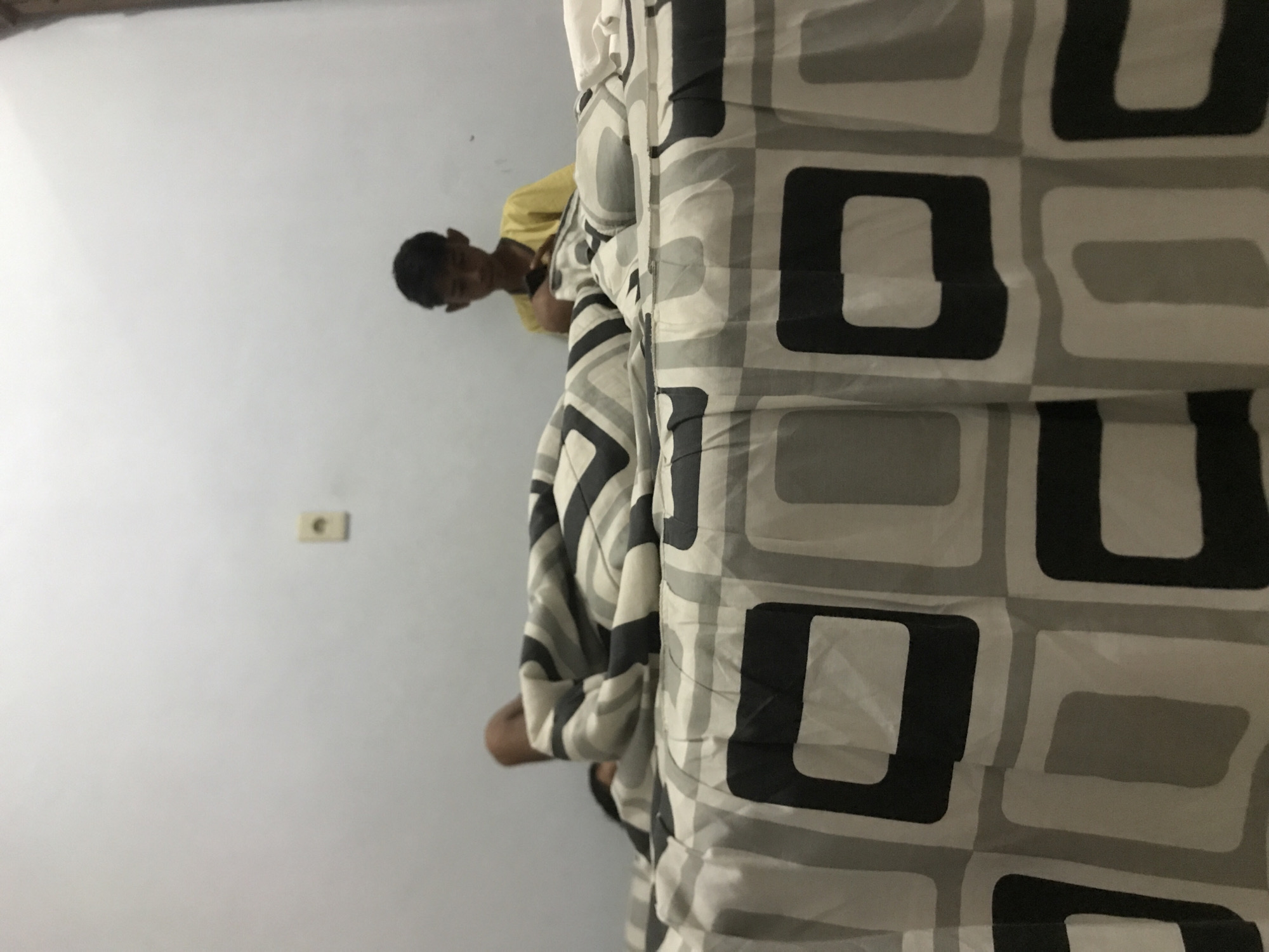 A low-angle indoor shot shows a young person in a yellow shirt standing partially behind a gray and black patterned comforter that dominates the right side of the frame. The person's face is visible, and they appear to be looking down. A wall is visible on the left side of the frame, featuring an electrical outlet. The location appears to be a bedroom, although the specific location within Lhokseumawe, Indonesia, is not discernible from the image.