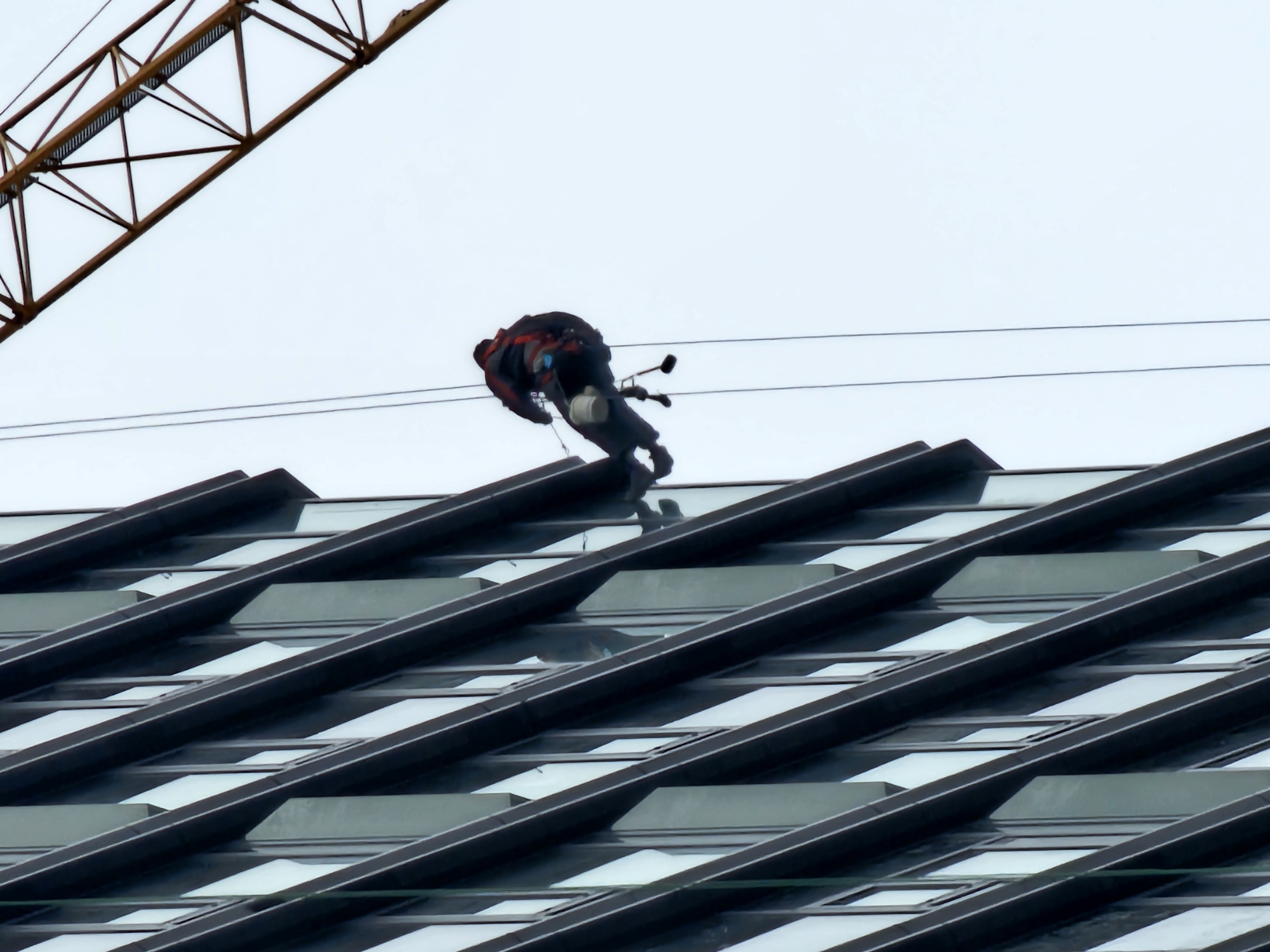 A high-angle outdoor shot captures a solitary worker rappelling down the exterior of a modern building in Moscow, Russia. The individual, likely a window cleaner or maintenance personnel, is suspended by several thin ropes or cables against a bright, overcast sky. They are dressed in dark clothing, possibly protective gear, with visible red straps or accents, and appear focused on their task, leaning forward with cleaning equipment such as a long-handled squeegee or brush and a bucket-like container attached to their harness.
In the upper left corner, the distinct yellow-orange lattice structure of a large crane boom is partially visible, indicating ongoing construction or industrial presence nearby. The building's facade, occupying the lower portion of the image, is characterized by a striking pattern of dark, diagonal architectural elements alternating with reflective or light-colored glass panels, creating a dynamic and layered effect. The scene takes place during the daytime under a uniformly bright, cloud-covered sky. There is no visible text in the image.