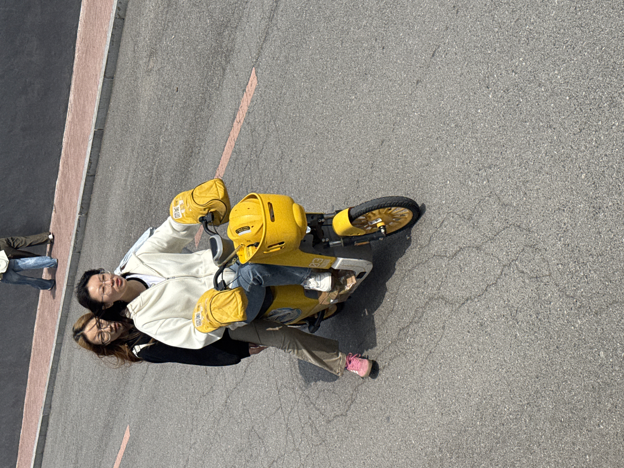 The image, captured from an elevated perspective and rotated, shows two individuals posing on a bright yellow electric scooter on an asphalt road in Zhongtun, China. Both subjects appear to be women, wearing light-colored hooded jackets and glasses, looking towards the camera. The person in the front has dark hair, wears blue jeans, and has yellow insulated covers or mittens over her hands on the handlebars. The person seated behind her has lighter brown hair, wears khaki pants, and pink sneakers.
The scooter is a prominent bright yellow, characteristic of delivery or shared e-bikes in China. Yellow bags are attached to the handlebars, bearing visible Chinese text. The text clearly reads "美团" (Meituan) and "买菜" (Maicai), identifying it as a Meituan Groceries delivery scooter. The number "03" is also visible on the scooter's body.
The scene is set on an asphalt road, showing some cracks and wear. A faded reddish-orange line is painted diagonally across the road. To the left, a sidewalk or curb with a reddish-brown strip is visible. The lighting is bright and clear, indicating a sunny daytime. In the background, partially visible, another person in a brown jacket and blue jeans walks away from the camera. The overall atmosphere is casual and relaxed, suggesting a moment of leisure or a photo opportunity.