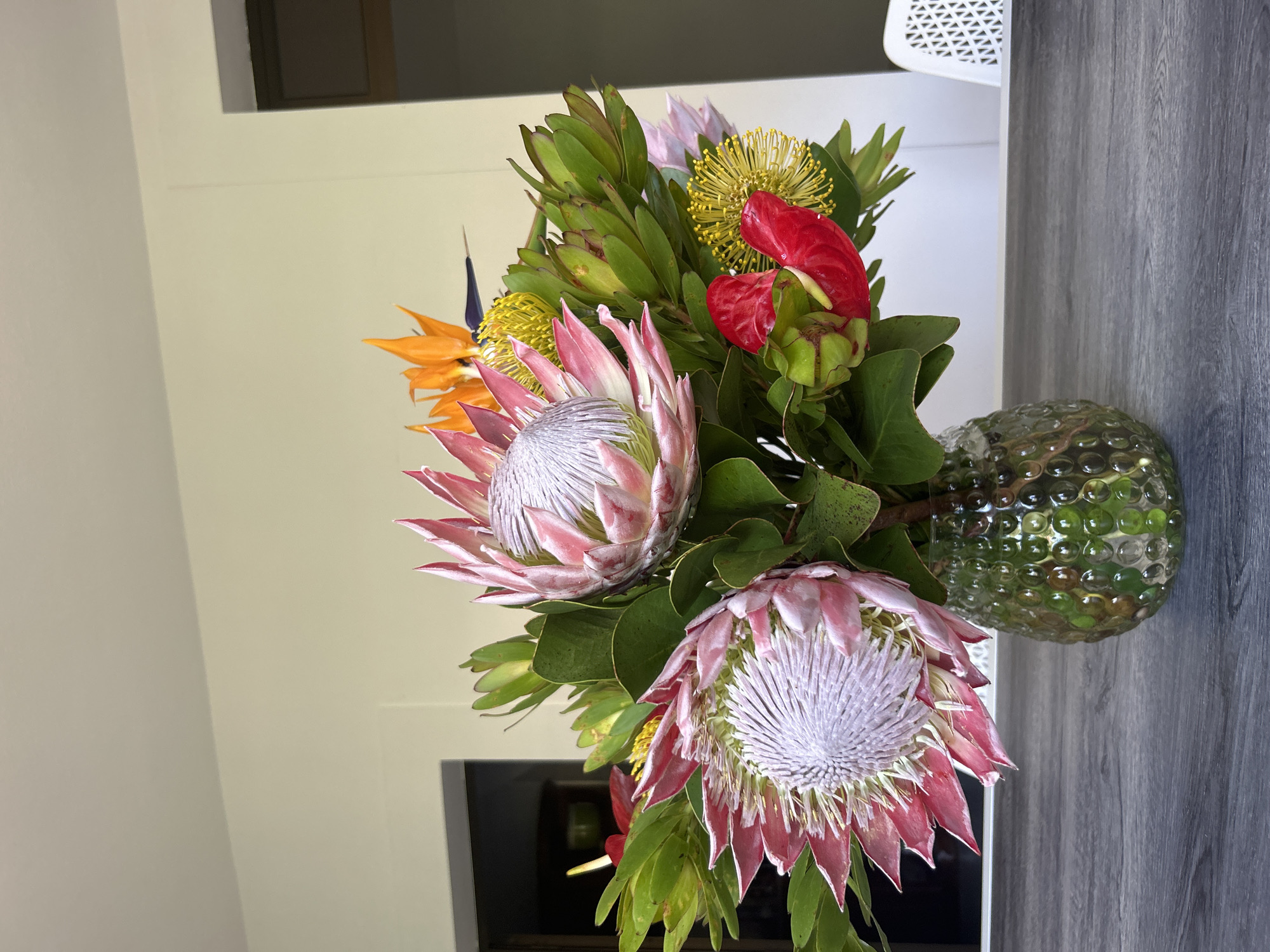 A vertical, medium-shot captures a floral arrangement in a speckled glass vase. The arrangement consists of two protea flowers with pink petals and white centers, accented by yellow pincushion flowers, an orange bird of paradise, and red anthurium flowers. Green foliage surrounds the flowers. The vase sits on a grey, wood-grain surface. The background consists of a light beige wall and a glimpse of a black object. The image does not appear to have been taken in Câmara de Lobos, Portugal; the setting is indoors and does not showcase features of the city.