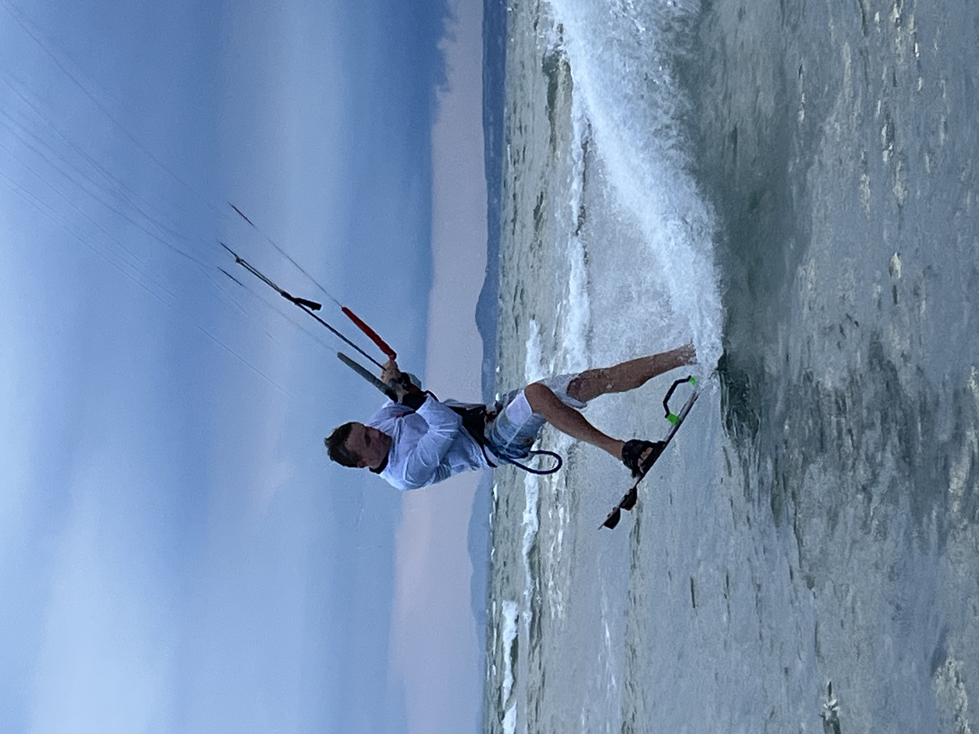 A person is kitesurfing on the water. The kitesurfer is a male wearing a white long-sleeved shirt, blue and white checkered shorts, and black sandals. He is holding onto a kite bar, which is attached to a kite in the sky (not fully visible). He is in mid-air, leaning back, with his board creating a spray of water. The water is choppy, with small waves. In the background, there are rolling hills and a cloudy sky. The scene appears to be set in Mũi Né, Vietnam, known for its water sports.