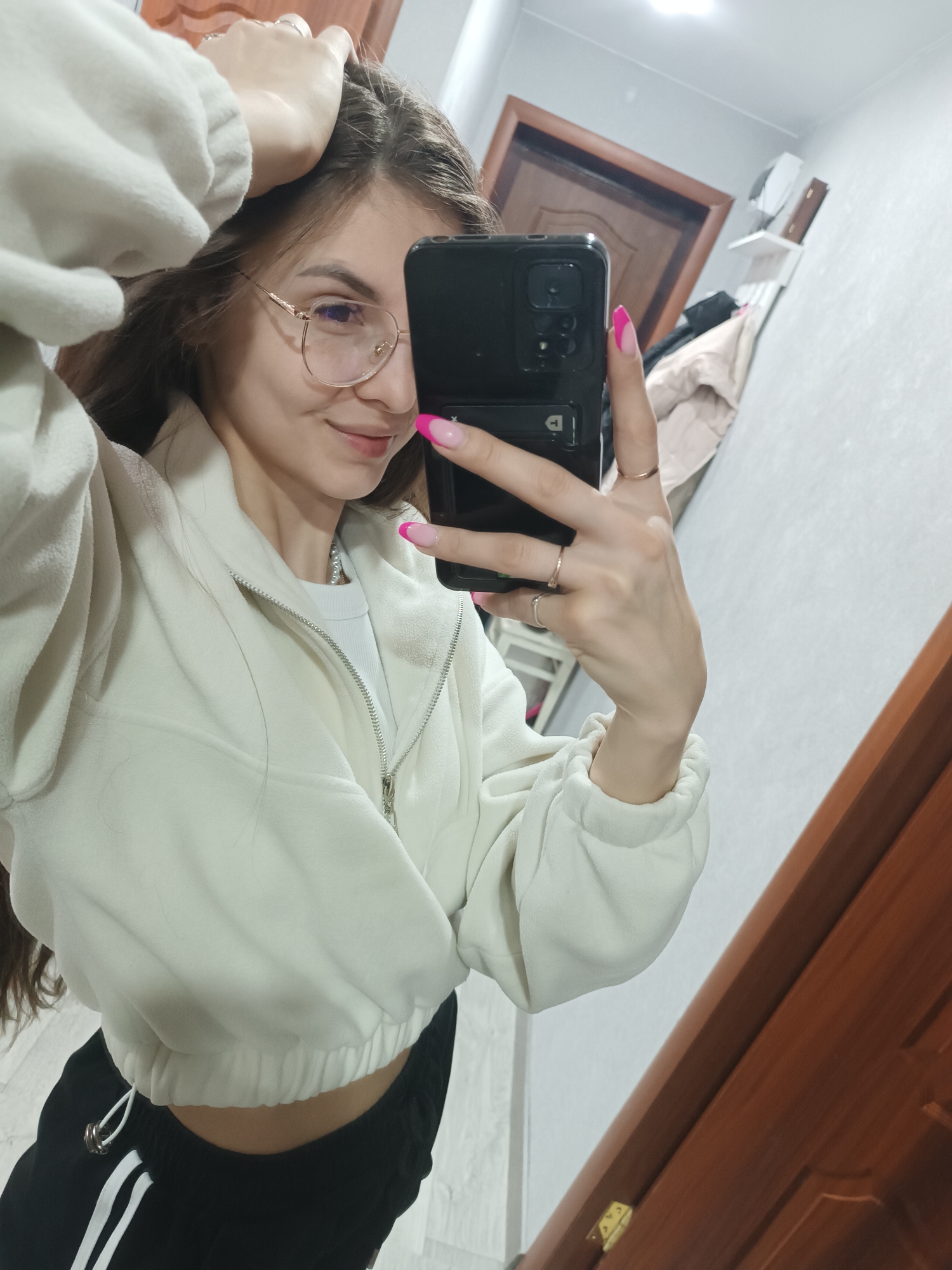 A female subject is performing a mirror selfie inside a room located in the city of Unknown, Unknown. The subject, positioned from the waist up, wears a white cropped fleece jacket, a white inner top, and black track pants with white vertical stripes. She has long brown hair, wears gold-rimmed eyeglasses, and exhibits a slight smile. A pearl necklace is visible. Her left hand, adorned with multiple rings and displaying pink and white French tip nails, holds a black smartphone vertically to capture the image. Her right hand is raised, with fingers positioned within her hair. The reflected background reveals light gray walls, a brown wooden door with a dark frame, a white ceiling light fixture, and a white wall shelf supporting two small objects. Below the shelf, a garment hangs. The floor appears light-colored and reflective.