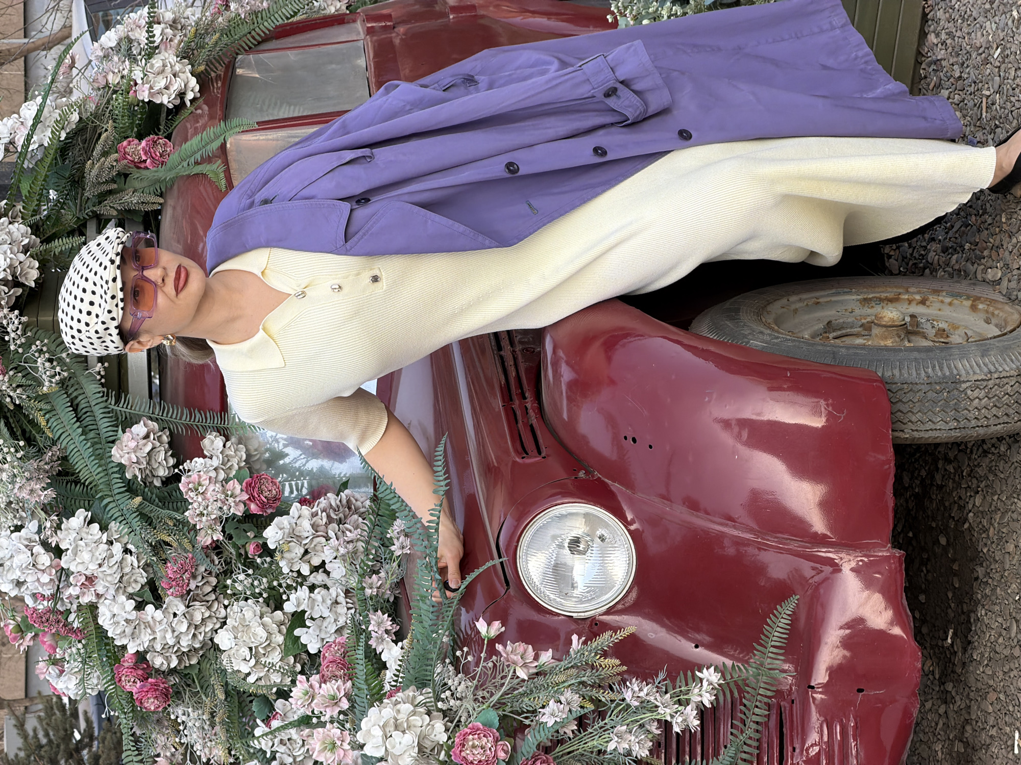 A stylish woman is captured in a full-body, posed shot, lying diagonally across the front of a vintage, deep red car. Her head is positioned to the left, looking directly at the camera.
The woman has a light complexion and is wearing a fashionable outfit. She sports a light yellow, ribbed, short-sleeved polo-neck top paired with flowing, wide-leg trousers or a skirt in a matching pale yellow or cream hue. A vibrant purple trench coat is draped over her torso and legs. Her head is covered by a white headscarf adorned with black polka dots, and she wears oversized purple-tinted square sunglasses and gold hoop earrings. Her lips are painted a dark red or berry shade.
The car, a vintage model in a glossy deep red or maroon color, forms the primary backdrop. Its prominent round headlight is visible on the lower right side. The car is extensively decorated with artificial floral arrangements, featuring large clusters of white, pink, and mauve hydrangea-like blossoms, small pink roses, and various green ferns and foliage. An old, rusty spare tire is visible leaning against the side of the car on the right.
The setting appears to be outdoors, with the ground beneath and around the car covered in gravel or small pebbles. The lighting suggests it is daytime. The woman's expression is neutral and composed, contributing to an overall chic and artistic mood. No visible text is present in the image.