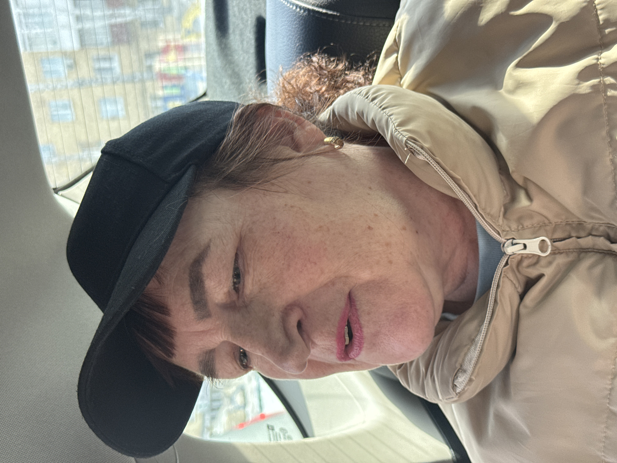 The image displays a woman seated in the rear passenger compartment of a vehicle, located in Votkinsk, Russia. The woman, appearing middle-aged, has fair skin with visible freckles and wrinkles, and dark brown hair with reddish highlights at the back. She wears a black baseball cap, a light blue undergarment, and a partially zipped beige puffer jacket. Her lips are accentuated with pink lipstick, and a gold-colored hoop earring is present in her right ear. She is positioned facing left within the frame, with her mouth slightly open, suggesting she is speaking or about to speak. Her eyes are directed forward or slightly downward.
Through the vehicle's rear window, a blurry urban background is visible. This includes a multi-story building with a light yellow facade and multiple windows, indicative of an apartment block. Below the building, indistinct, colorful shapes (red, blue, yellow, green) are discernible, consistent with elements of a children's playground. The interior of the vehicle shows a light grey headliner and a dark grey or black headrest from the seat. Daylight illuminates the scene.