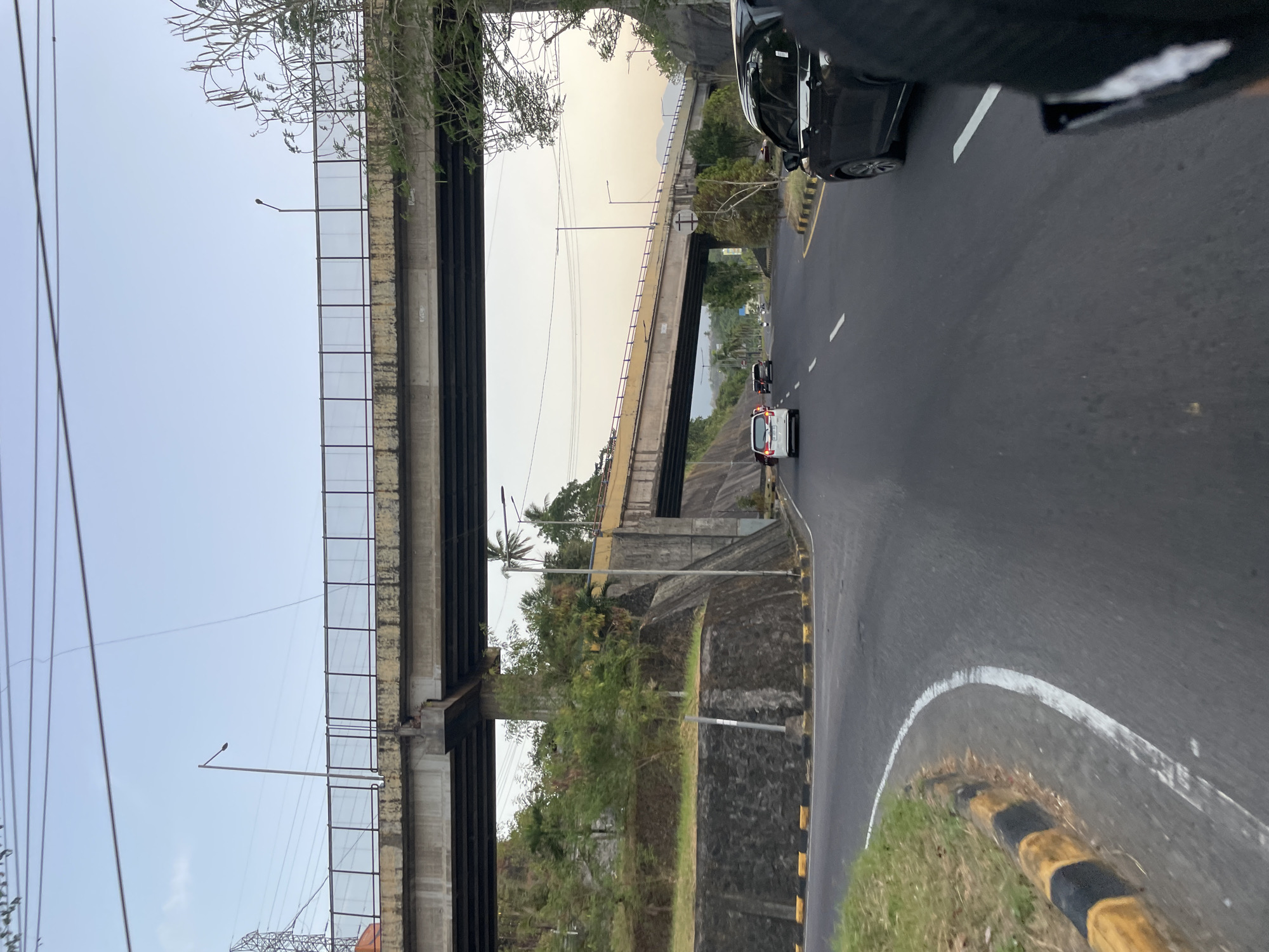 The image depicts a road with two vehicles and a pedestrian overpass in Maumbi, Indonesia. The overpass, constructed of concrete and metal railings, spans over the road. A street light is visible on top of the overpass. The road itself is paved and marked with white lines, and features a grassy embankment on the left side with yellow and black striped barriers. On the road, a black car is seen in the foreground on the right, and a white car is in the distance on the left, along with two other vehicles and a motorcycle. The scene is set against a backdrop of trees and hills, under a pale sky. The overall impression is that of a typical urban or suburban transportation artery.