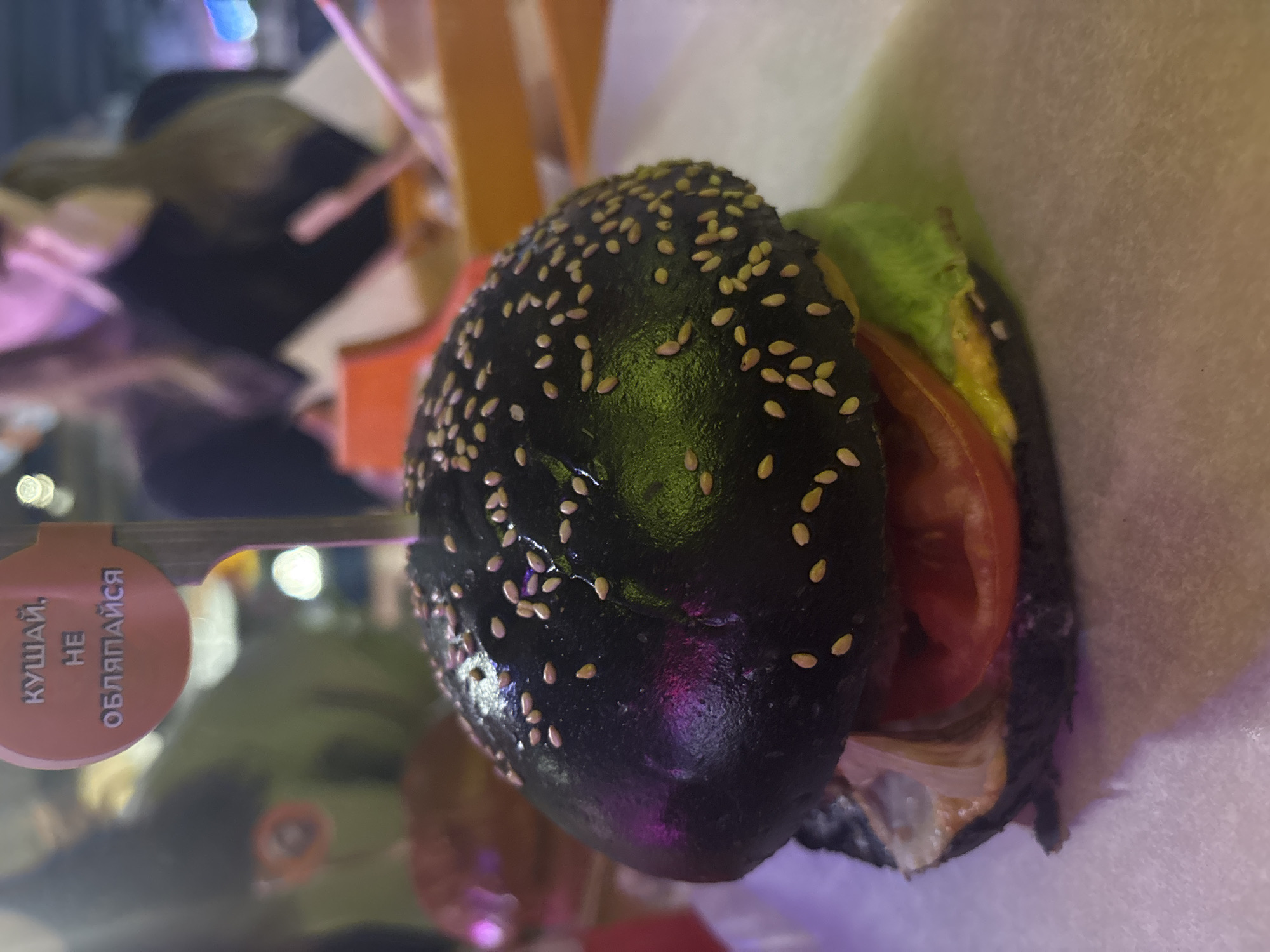 A black sesame seed bun burger with tomato, lettuce, and sauce filling is pierced with a skewer bearing a pink tag with the text "КУШАЙ, НЕ ОБЛЯПАЙСЯ" (EAT, DON'T GET DIRTY). The location is likely a restaurant or food stall in Saint Petersburg, Russia, considering the Russian text on the tag. The burger sits on a white surface and appears to be indoors under artificial lighting.