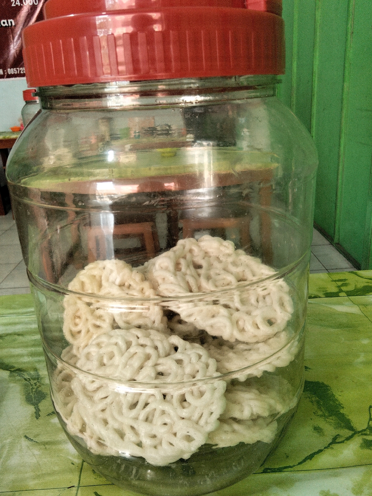 A large, clear glass jar with a red lid stands on a green, patterned surface. The jar is filled with numerous white, circular, and intricately shaped crackers, stacked on top of each other. The crackers have a layered, airy appearance. The background of the image shows a green, vertical surface, possibly a wall, with a hint of a table and chairs reflected in the glass jar. The setting appears to be an outdoor or semi-outdoor location, possibly a food stall or a market in Panggang, Indonesia. No people are visible in the image.