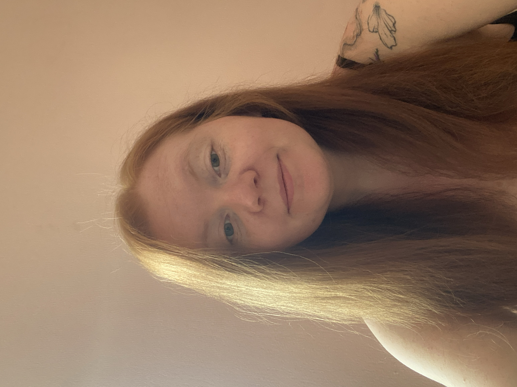 A close-up portrait shows a fair-skinned person with light reddish-blonde hair, possibly female, smiling slightly. The background is a plain beige wall. A tattoo is visible on the upper arm. The location is not discernible from the provided image; there is no indication that this photo was taken in Yekaterinburg, Russia, or any other specific location.