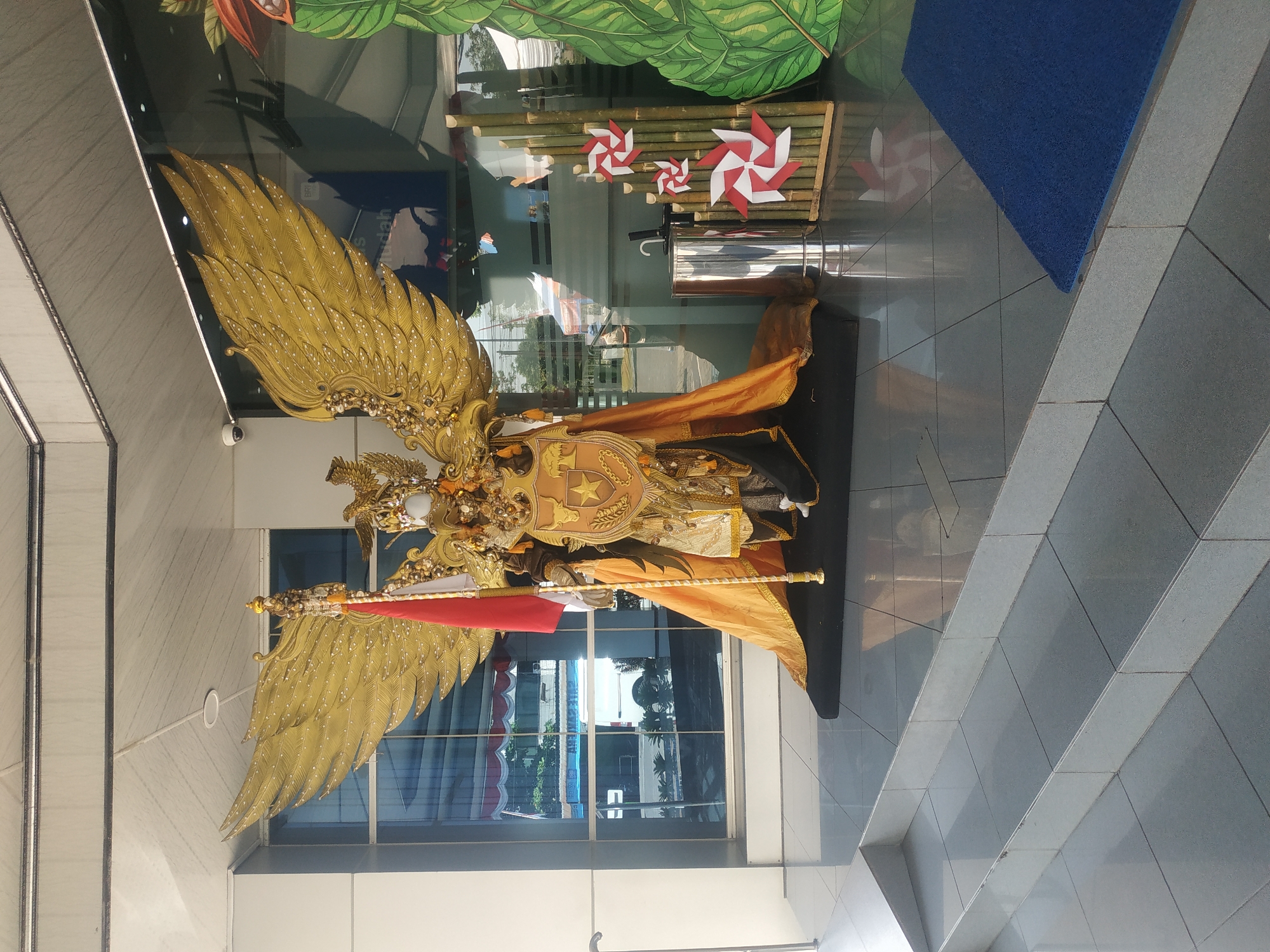 The image shows a large, ornate costume resembling Garuda, a mythical bird from Indonesian folklore, standing on a raised platform in front of a building with large glass windows. The costume is gold and adorned with intricate details, featuring large wings, a shield with a star emblem, and a golden cape. A red and white Indonesian flag is mounted on a staff next to the costume. To the right of the costume, there is a bamboo display with red and white pinwheel decorations and a large green leaf cutout. A metal trash can is also visible. The building appears to be a modern office or public building, with reflections of the outside environment visible in the glass. The scene is set outdoors on a sunny day, with stairs leading up to the platform. The location is identified as Jember, Indonesia.