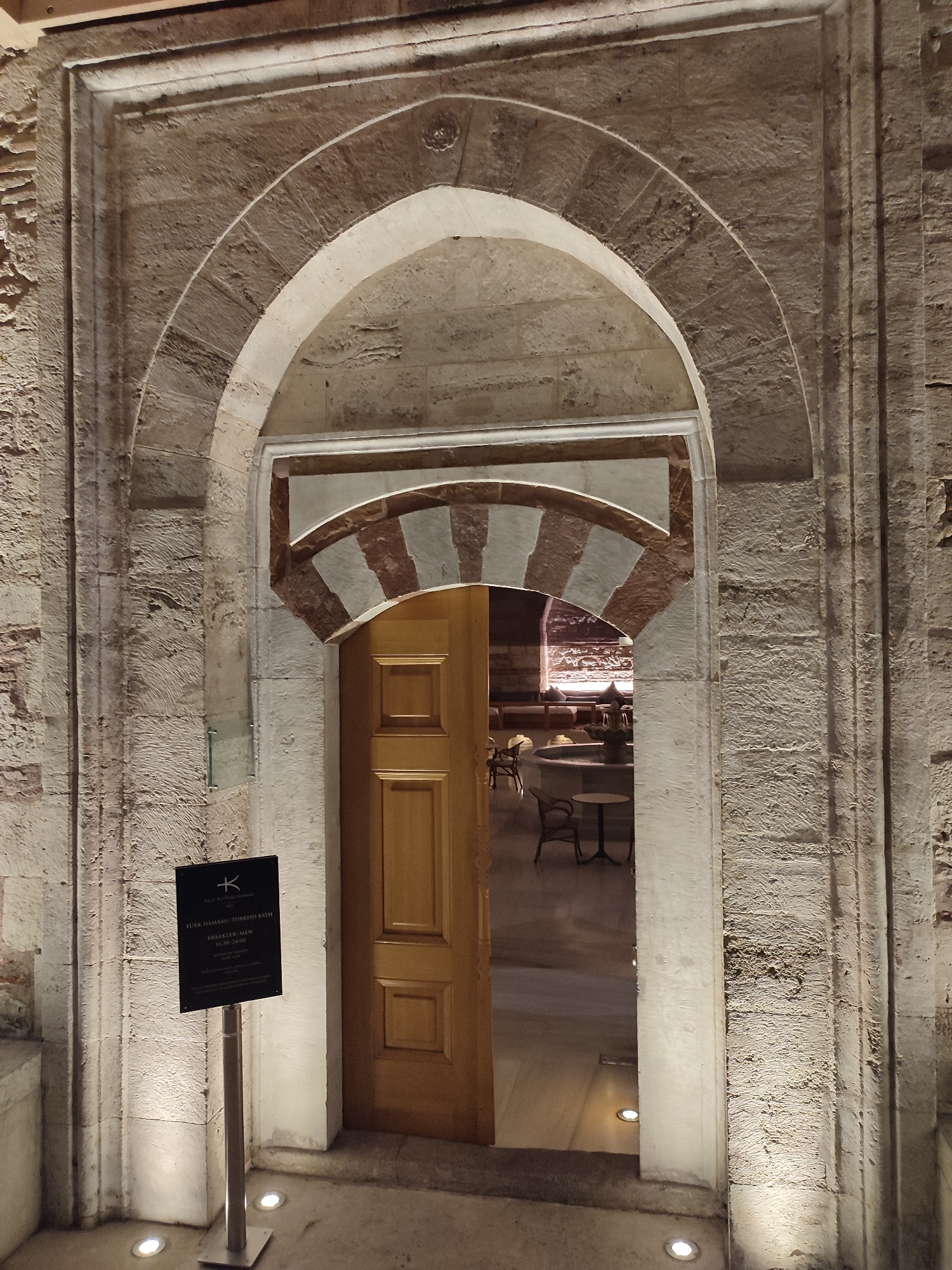 The image shows an arched stone entryway to a building. The entryway features rustic stone walls and a decorative arch with alternating light and dark stone segments. A wooden door is partially open, revealing a glimpse of an indoor seating area with tables and chairs. A black sign on a metal pole stands to the left of the doorway. The scene is illuminated by spotlights at the base of the entryway and from within the building. The architecture suggests a historical setting. Based on the sign that reads "Türk Hamamı" (Turkish Bath) and its stylistic features, this entryway is likely located in the Beyoğlu district of Istanbul, Turkey, possibly leading to a traditional hammam.