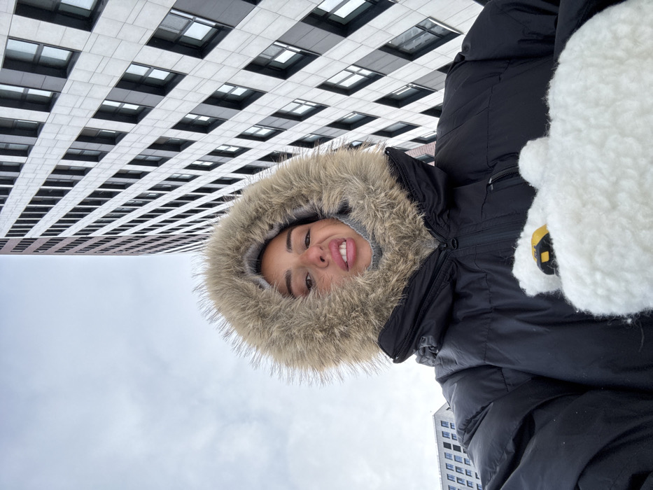 A female subject, centered in a low-angle selfie, is depicted outdoors in Moscow, Russia. She wears a black winter parka with a large, light brown fur-trimmed hood and a grey balaclava or neck gaiter covering her lower face. Her lips are formed into a pout, and her nose is slightly crinkled. Her right hand, partially visible, is covered by a white, textured mitten, from which a yellow and black tape measure protrudes. In the background, a tall, modern high-rise building dominates the right side of the frame, featuring a facade of light grey rectangular panels alternating with dark, recessed rectangular windows. A smaller, light-colored modern building is partially visible on the far left. The sky is uniformly overcast and light grey. The architecture of the prominent building is consistent with the Headliner Residential Complex (ЖК "Хедлайнер") located on Shelepikhinskaya Embankment, Presnensky District, Moscow.