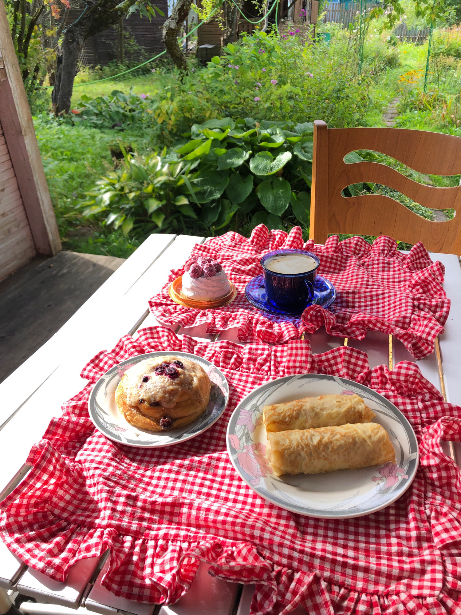 A white picnic table is set with breakfast items and a blue cup of coffee, arranged on a red and white checkered cloth with ruffles. On the table, there are two rolled pancakes on a floral-patterned plate, a pastry topped with raspberries on a gold pedestal, and a single round pastry with visible berries and icing. In the background, a wooden chair is visible, and beyond that, a lush green garden with various plants and trees, suggesting an outdoor setting in a rural or suburban area, possibly a dacha or garden society. The sunlight indicates it is daytime. The scene depicts a peaceful morning meal outdoors.