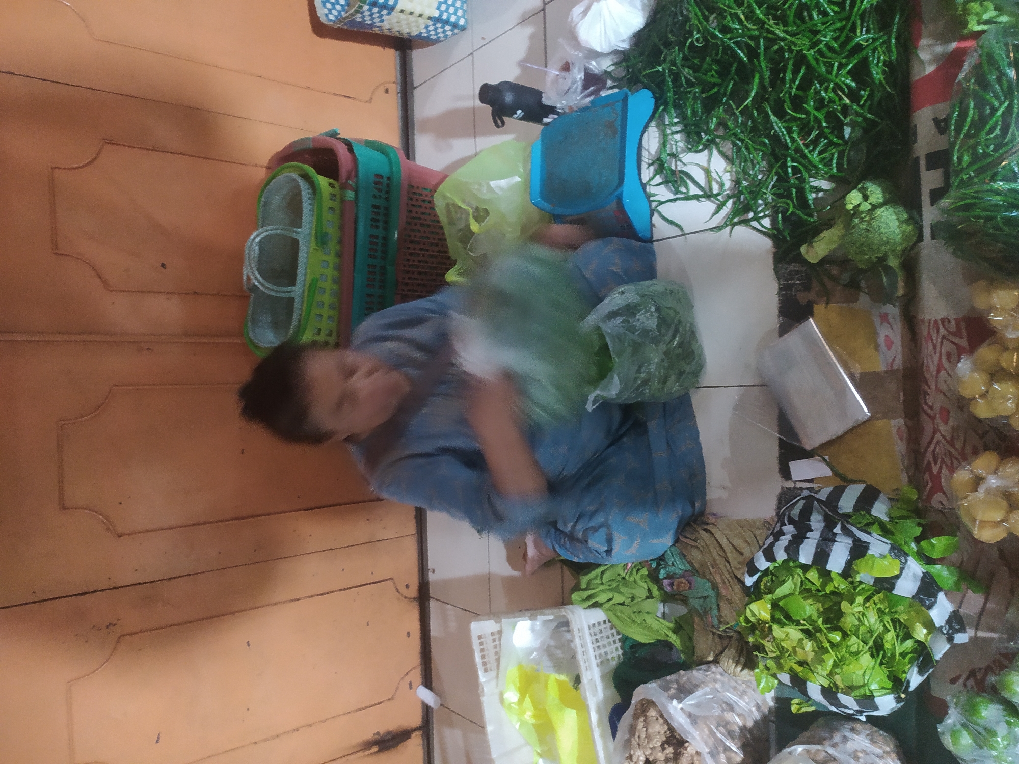 An individual is seated on the floor in front of a closed, wooden door, surrounded by various produce and market items. The individual, wearing a blue patterned garment, is in the process of bagging leafy greens. In the background, several plastic baskets are stacked, one with a green and white pattern, another predominantly green. To the right of the individual, a blue weighing scale is visible, along with a black water bottle and a clear cup with a red liquid. In front of the individual, there are bags of green chilies, broccoli, and other assorted vegetables. The floor appears to be tiled. The scene suggests a market or home-based food stall, possibly in the city of Kupang, Indonesia.