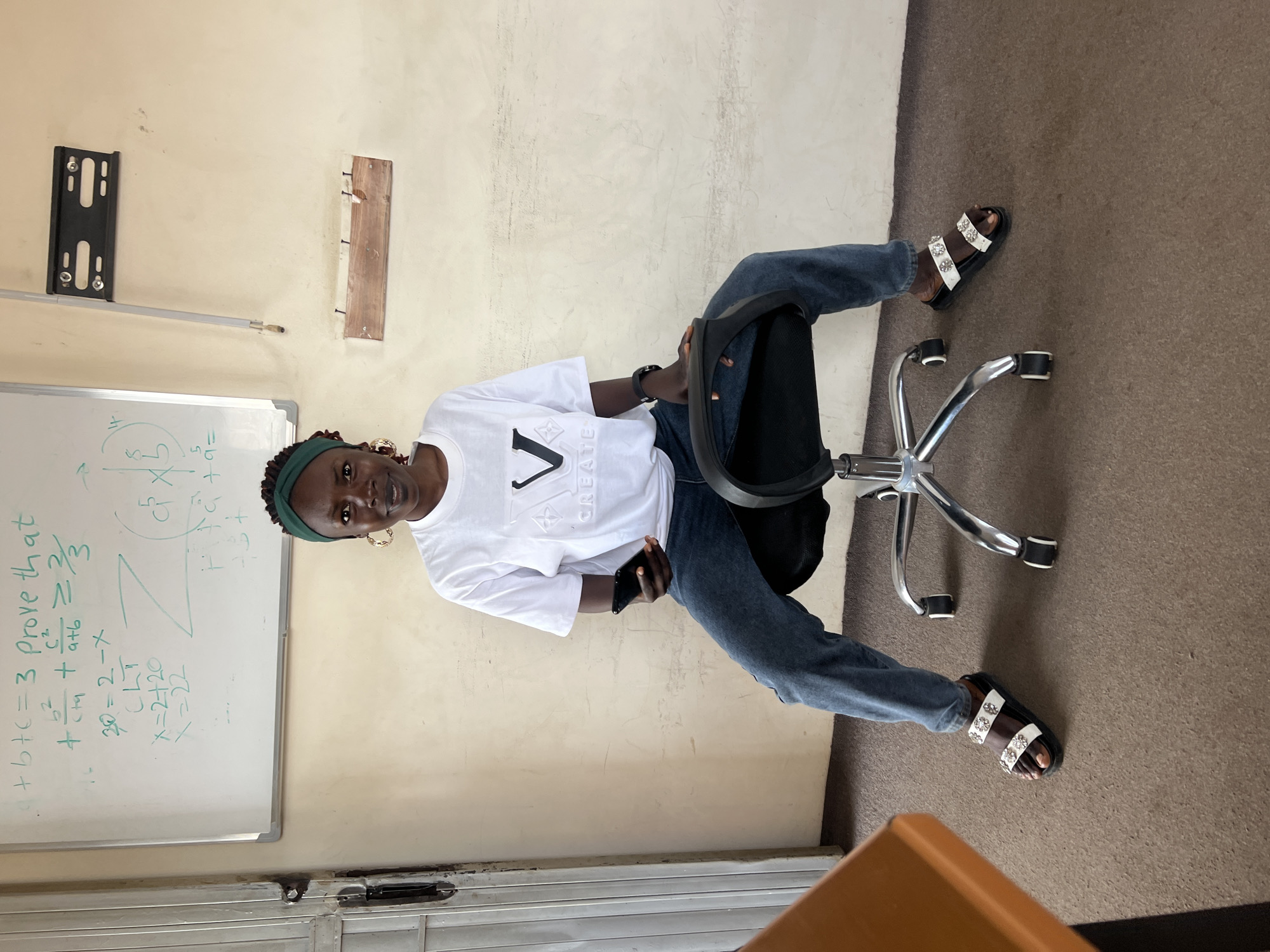 A young Black woman sits on a rolling office chair in front of a whiteboard covered in mathematical equations. She is wearing a white t-shirt with a black logo, blue jeans, and white sandals adorned with rhinestones. A green headband is in her hair, and she is holding a black smartphone in her right hand. The whiteboard, mounted on a wall with textured plaster, displays complex equations and diagrams. To the left of the whiteboard, a metal door is visible. To the right of the whiteboard, a wall-mounted bracket and a piece of wood with nails are attached. The floor is covered in brown carpet. The scene appears to be an indoor classroom or study area.