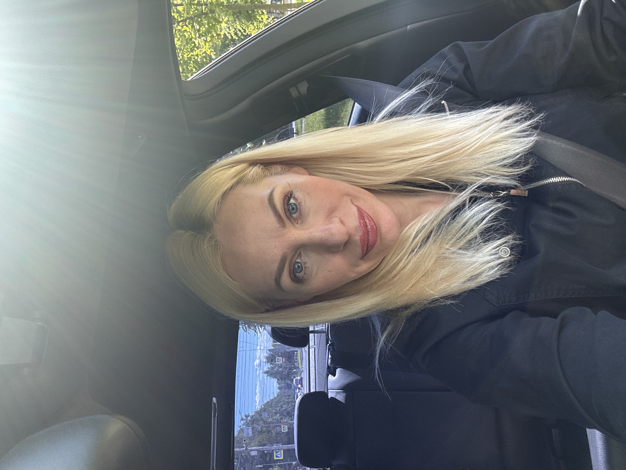 A woman with blonde hair is taking a selfie in a car. She is wearing a black jacket and has on makeup. Her hair is partially covering her face. The sun is shining brightly, creating lens flare. Outside the car window, trees and buildings are visible, suggesting an urban or suburban setting. The image appears to be taken during the daytime. The background shows a street scene with traffic signals and some foliage.